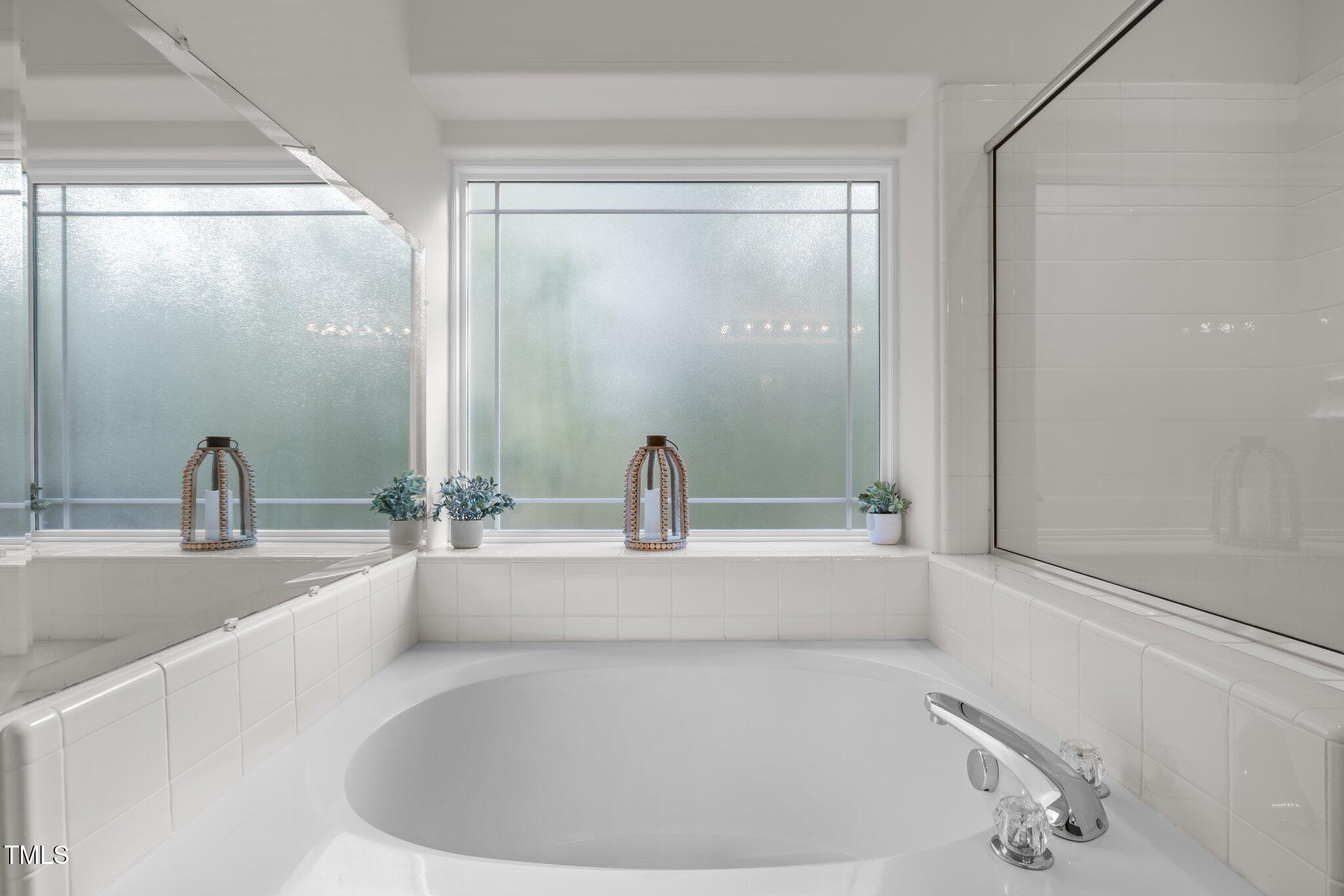 Built-in soaking tub beneath frosted windows, with white tile surround and decorative plants.