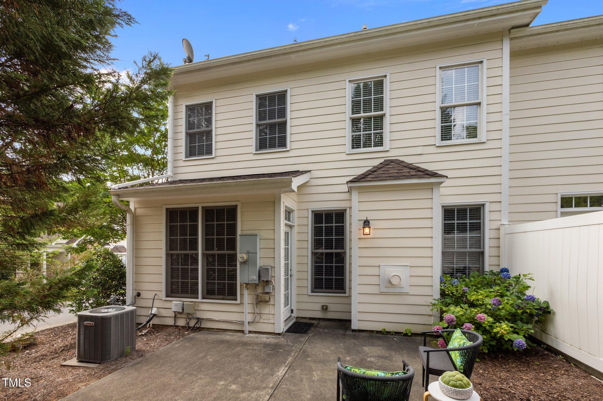Rear of a two-story beige home with a paved patio, outdoor chairs, and hydrangeas beside a white privacy fence.