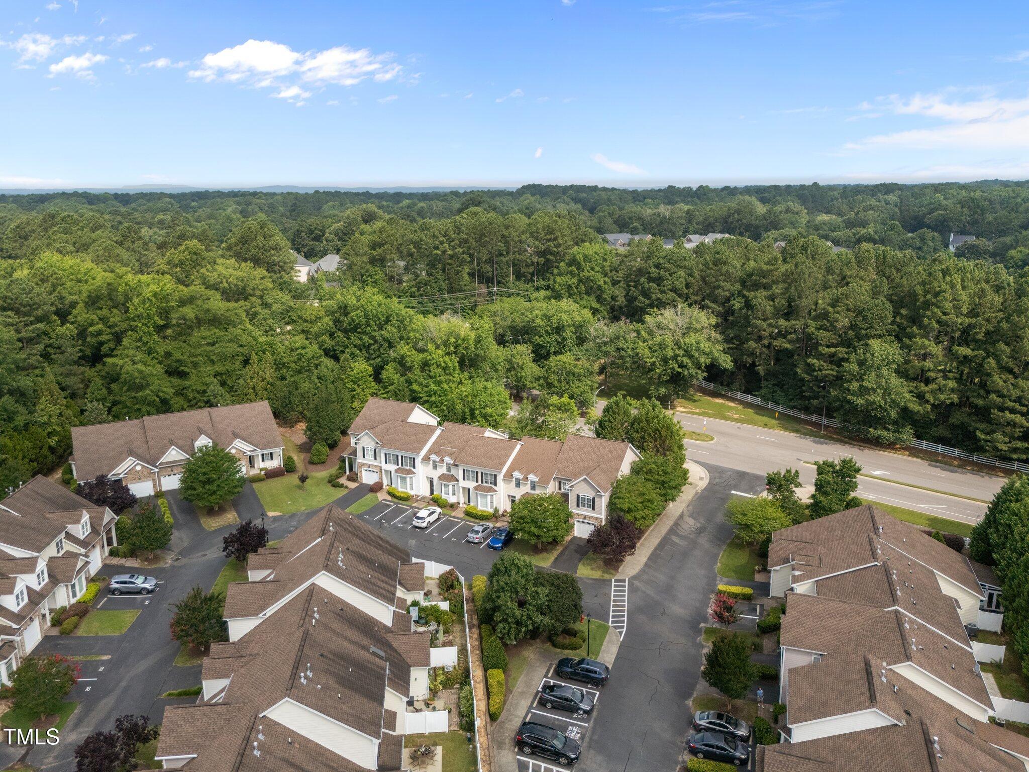 Aerial view of attached townhomes beside a tree-lined road and surrounding woodland.