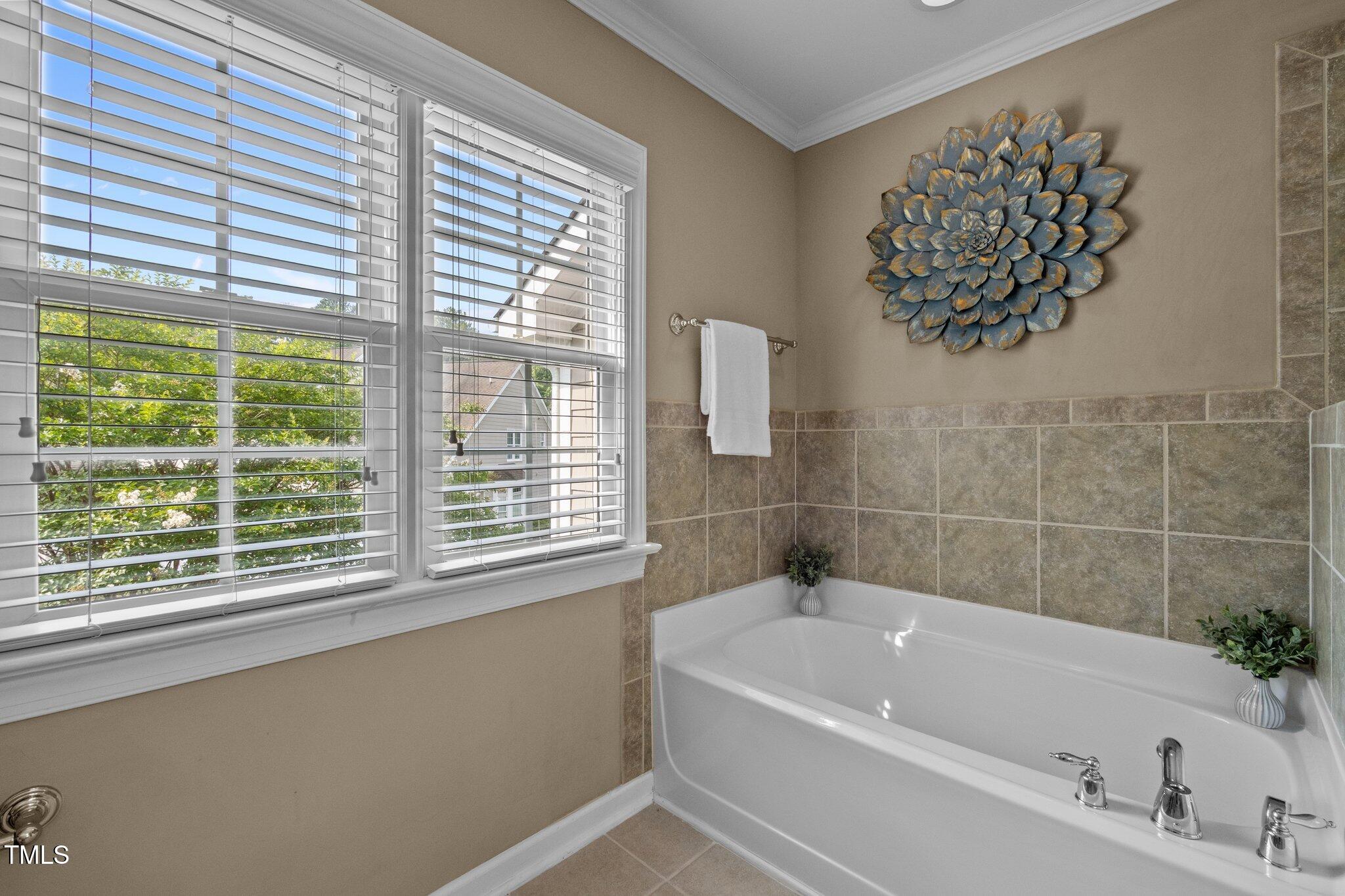 Bathroom with a white bathtub, beige tile surround, and two large windows with blinds.