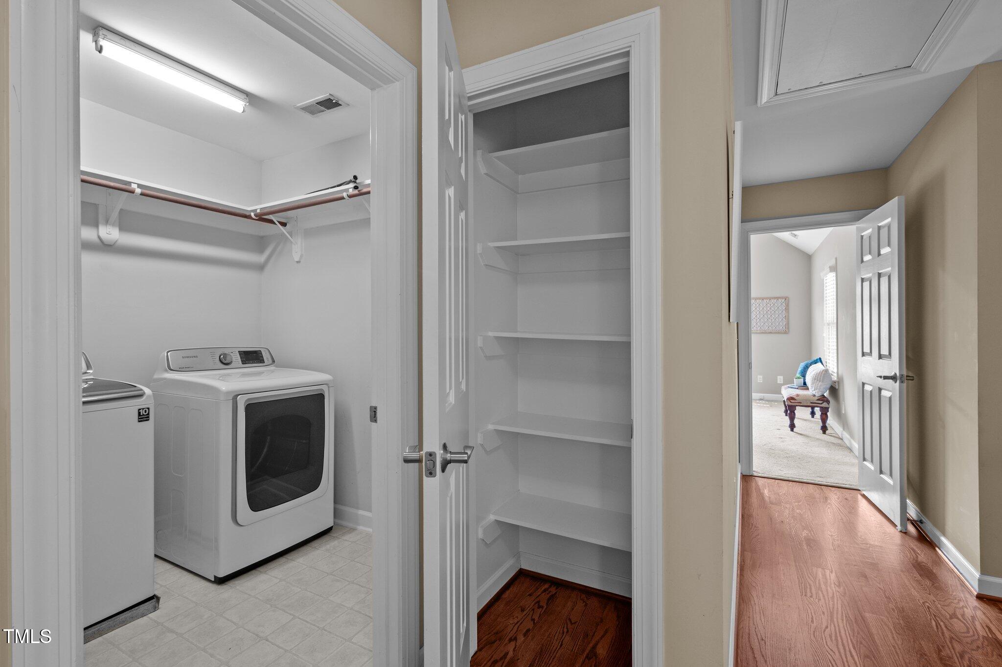 Laundry room with washer and dryer beside a tall shelved closet and hardwood hallway.