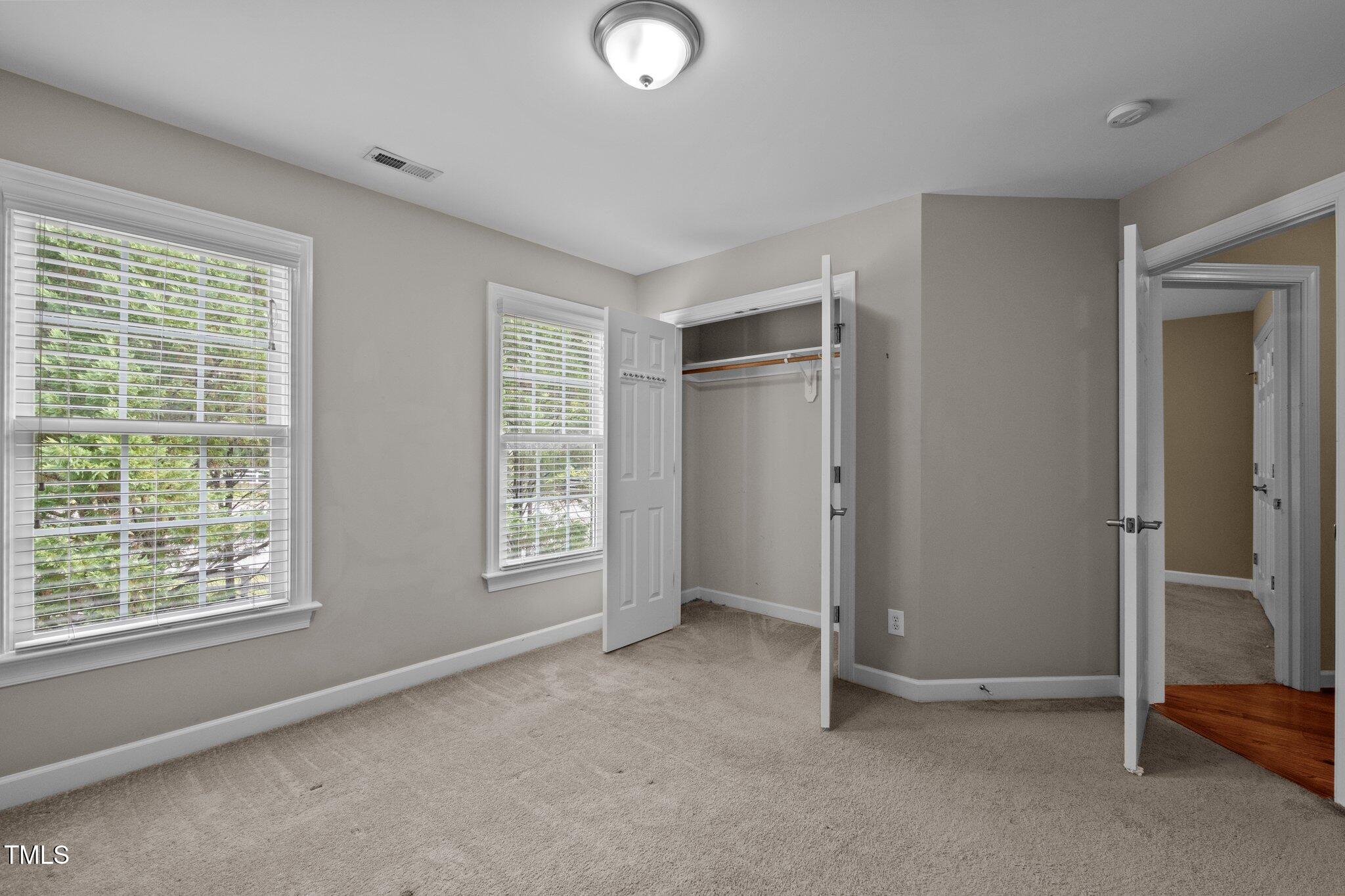 Empty carpeted bedroom with an open closet and two windows with white blinds.