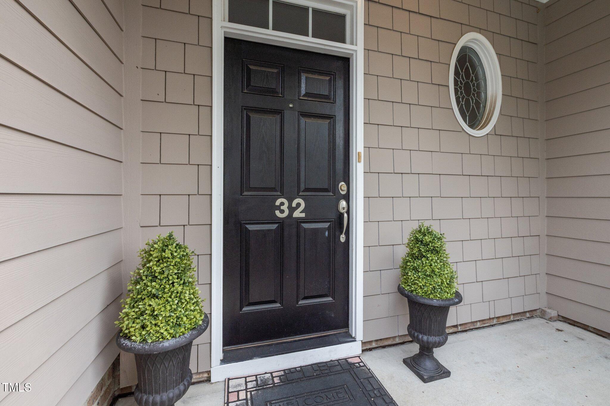 Unit 32 entrance with a black paneled door, oval window, and potted shrubs.