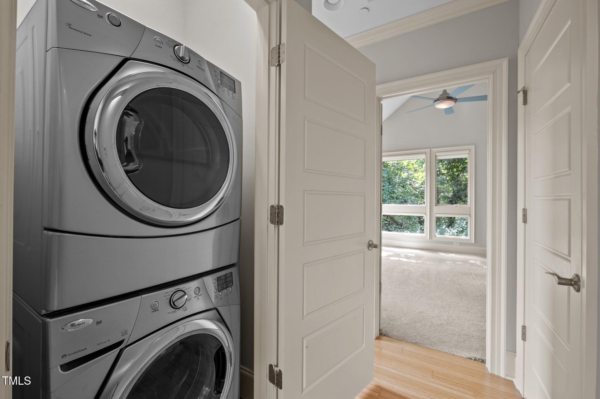 Stacked washer and dryer beside an open doorway to a bright, carpeted bedroom.