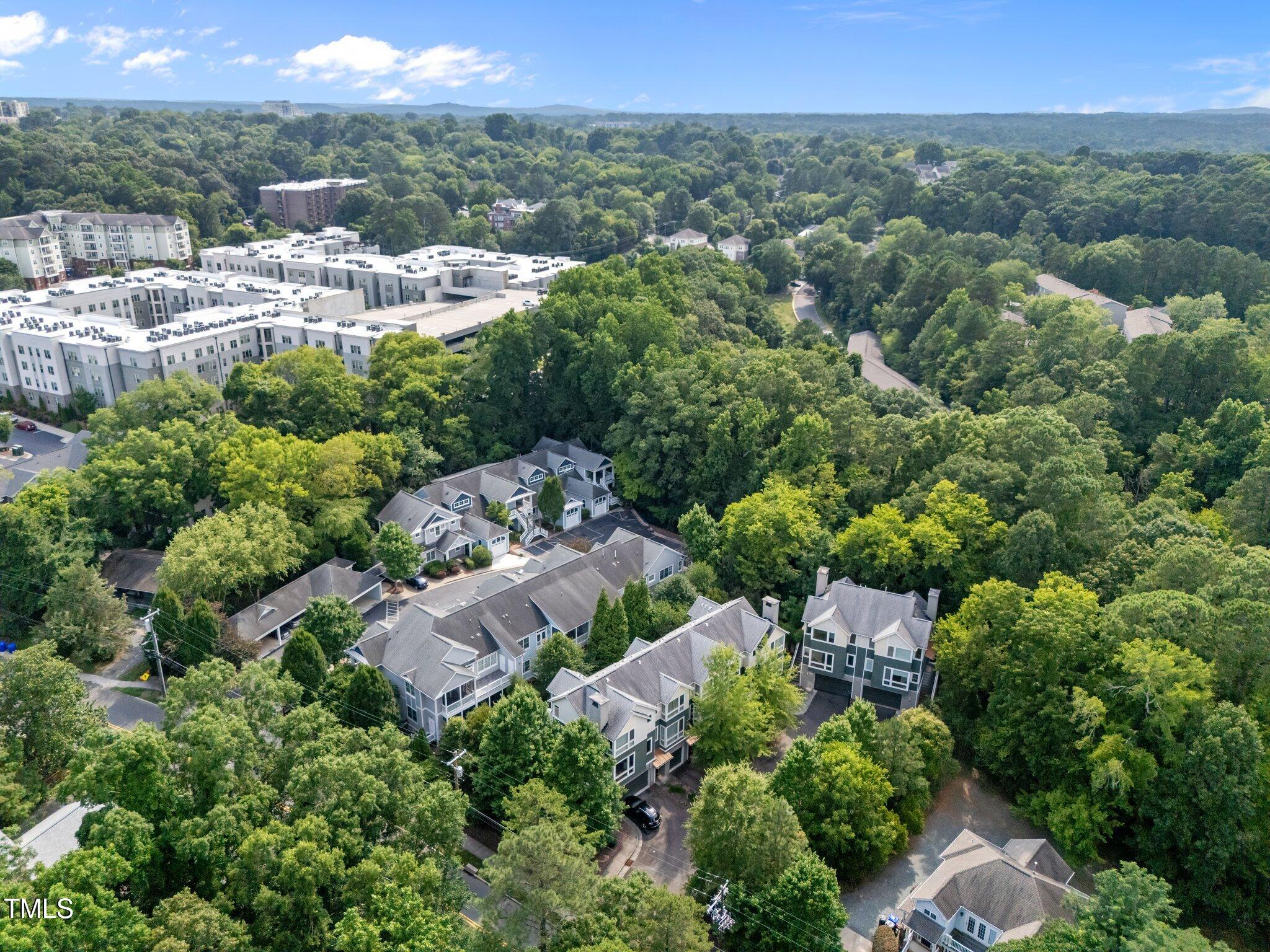 Aerial view of gray-roofed residences surrounded by trees, with larger apartment buildings in the distance.
