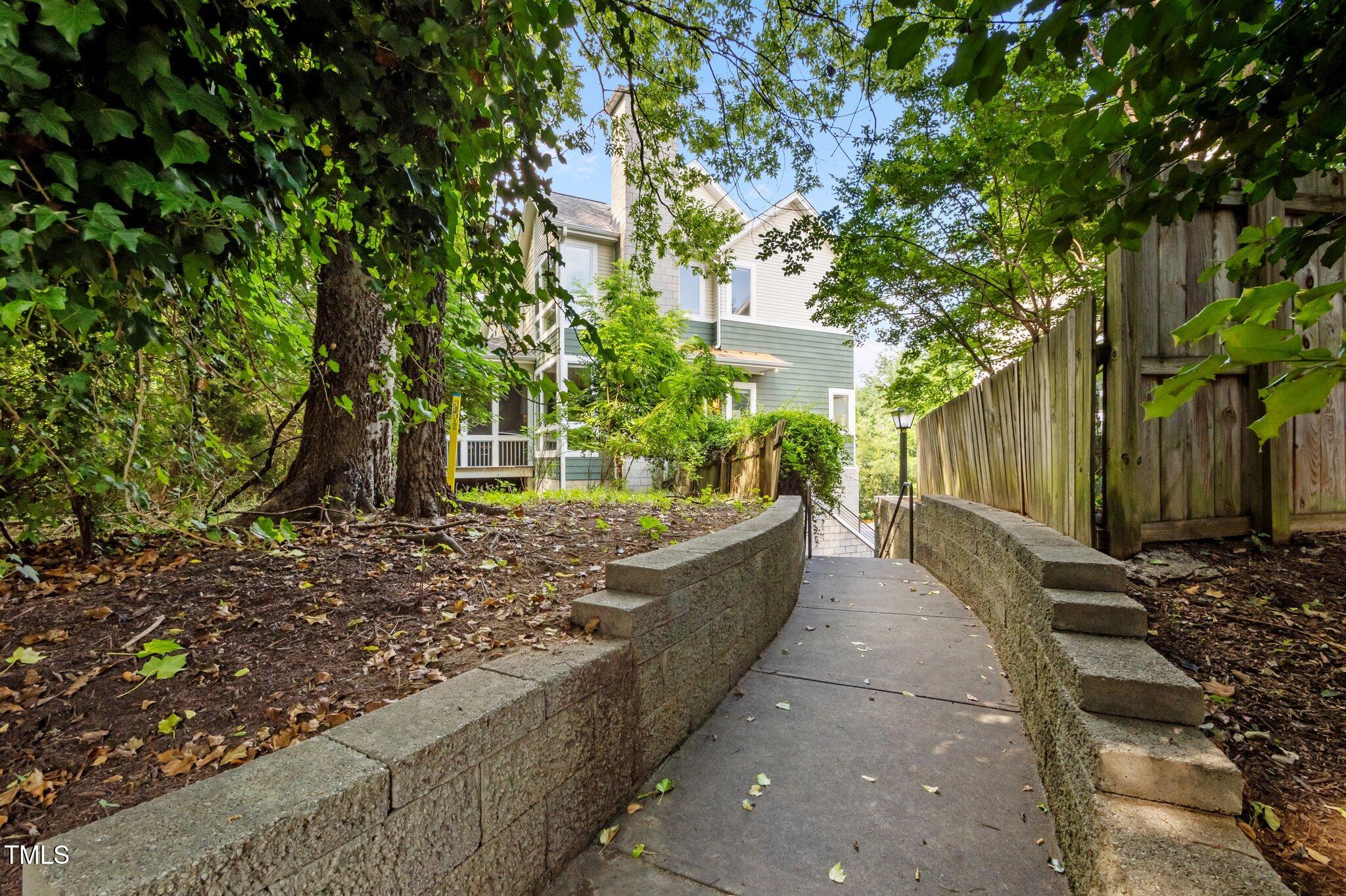 Walkway to a multi-story home through leafy landscaping, between stone retaining walls and a wooden fence.