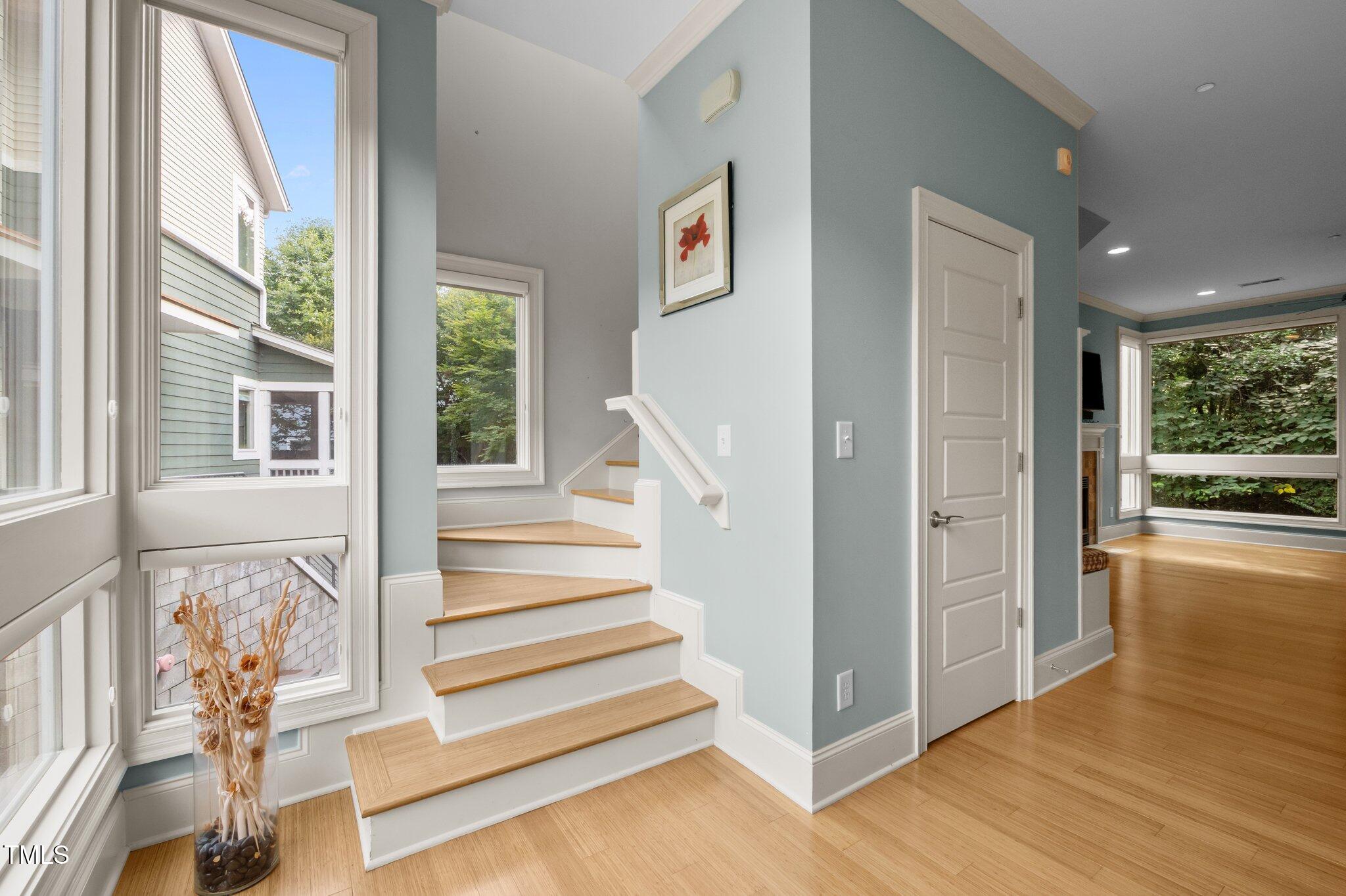 Interior staircase with light wood steps beside large windows overlooking trees and an adjoining hardwood-floored room.