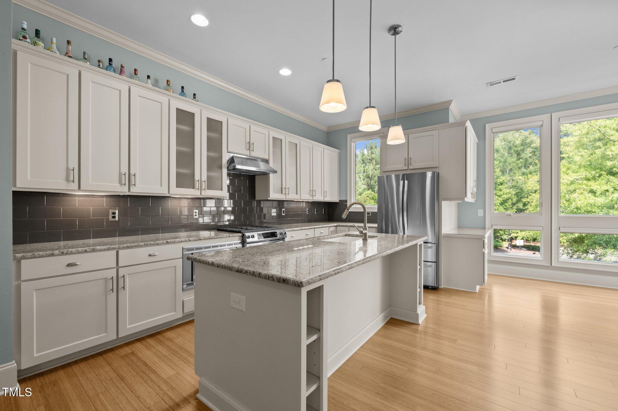 Kitchen with a granite island, stainless steel appliances, dark tile backsplash, and large windows.