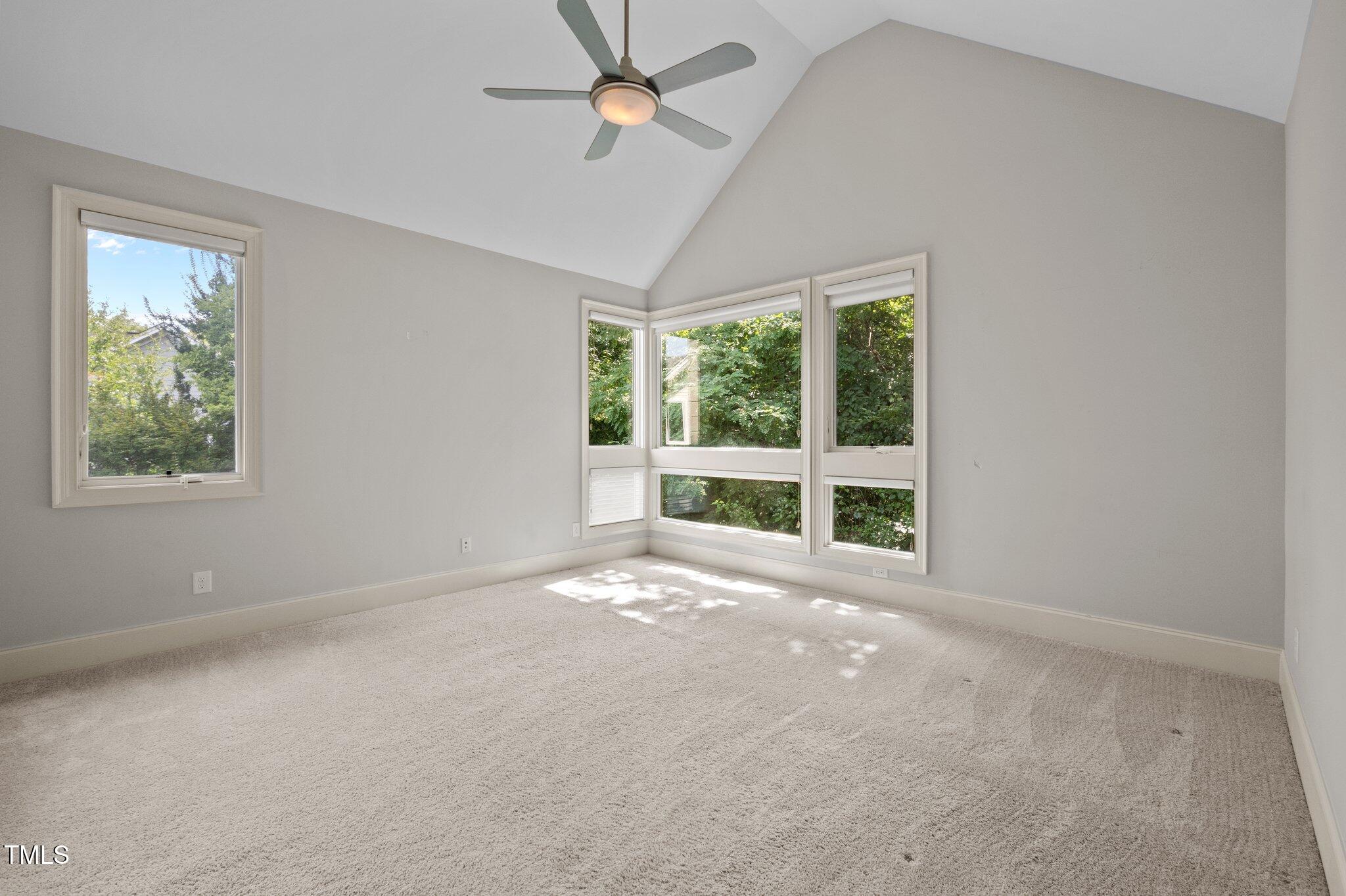 Vacant bedroom with a vaulted ceiling, ceiling fan, and corner windows overlooking greenery.