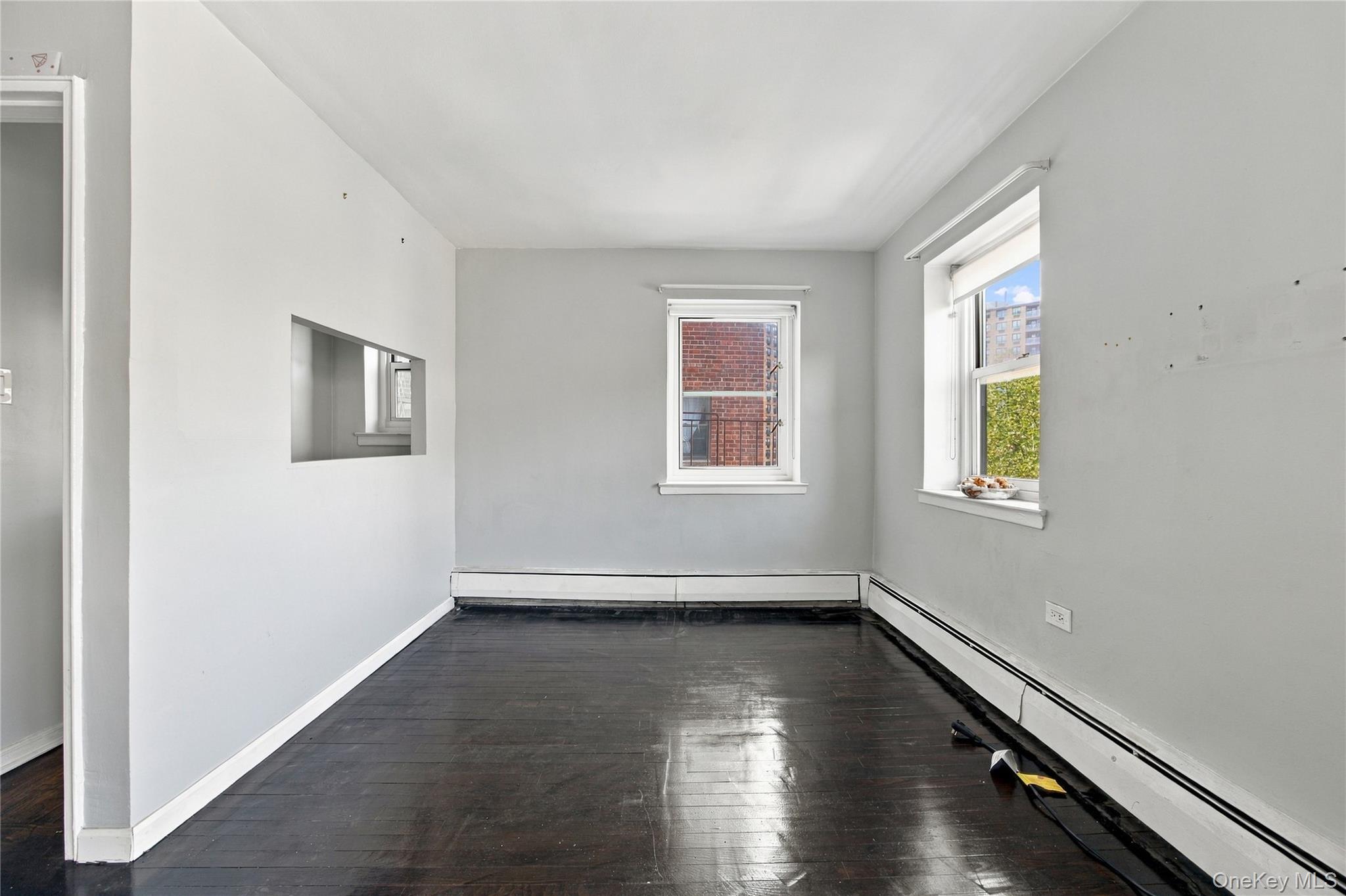 Empty room with dark hardwood floors, two windows, and baseboard heaters.