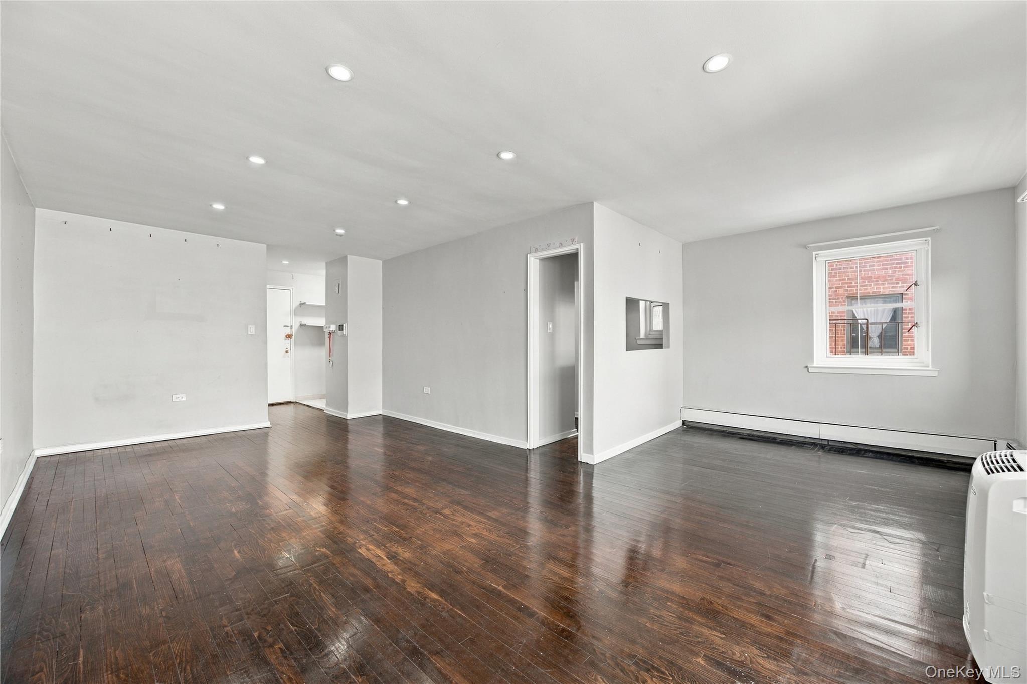 Vacant living room with dark hardwood floors, recessed lights, and a window overlooking a brick building.