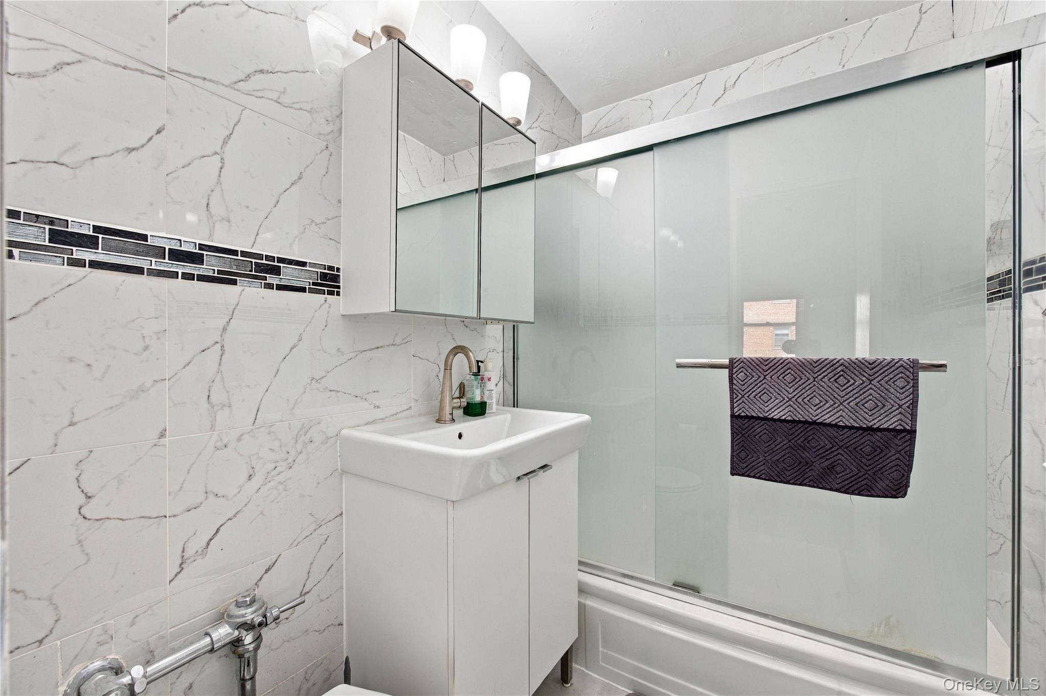 Bathroom with marble-look tile, a white vanity, and frosted glass tub-shower enclosure.