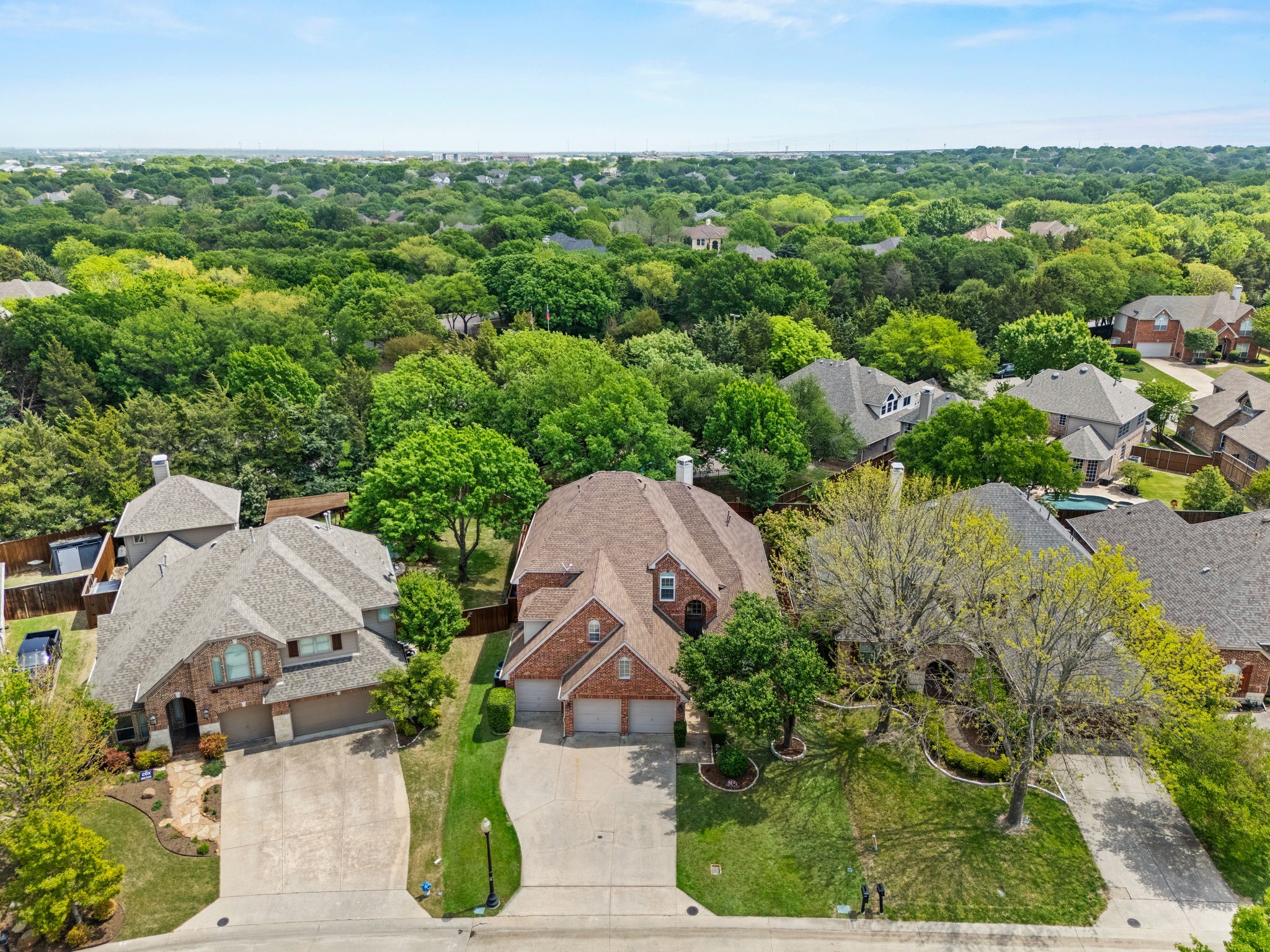 Aerial view of a red-brick house and driveway among mature trees and neighboring homes.