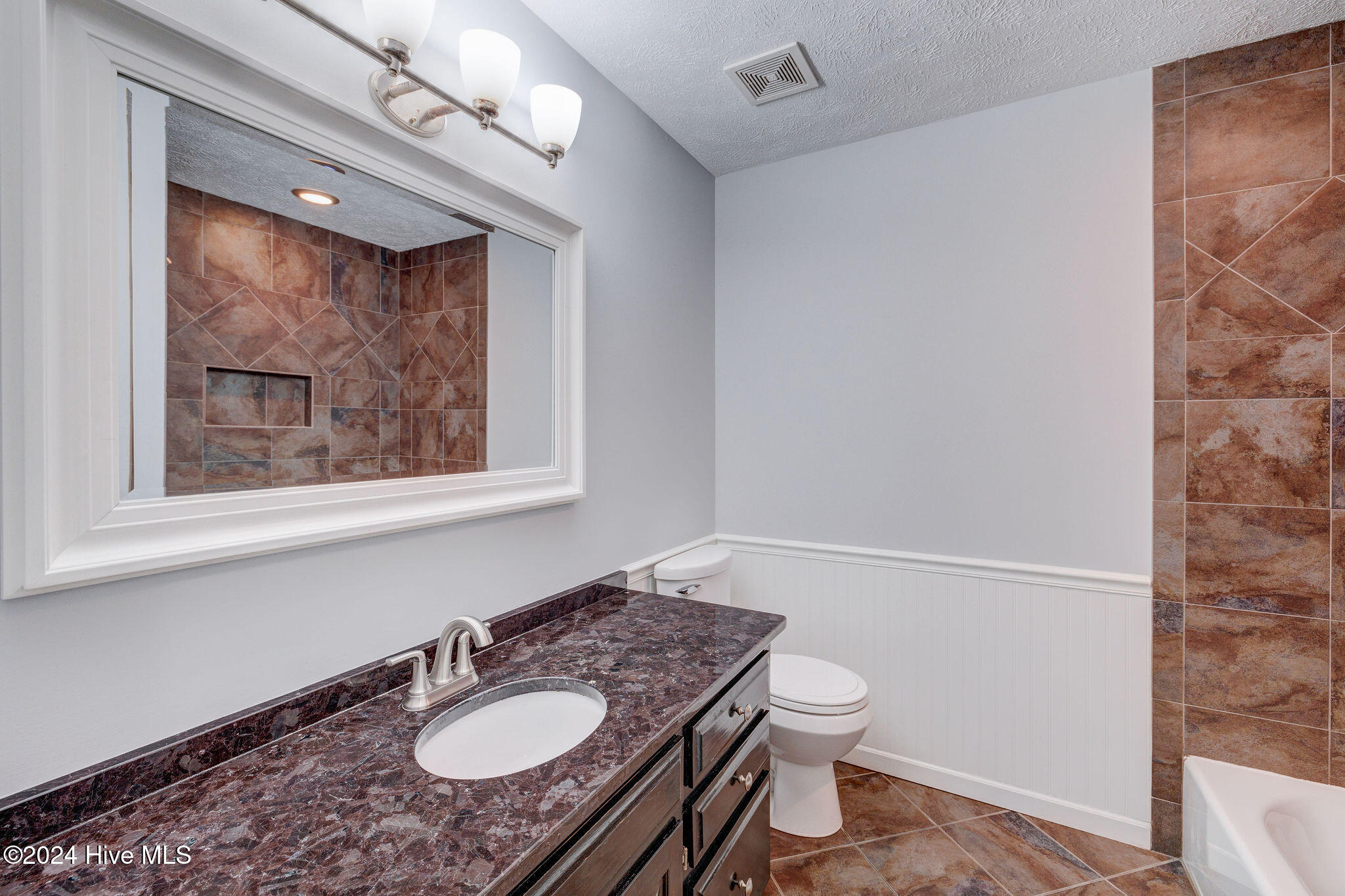Bathroom with a dark stone vanity, large mirror, brown tiled shower wall, and white paneled wainscoting.
