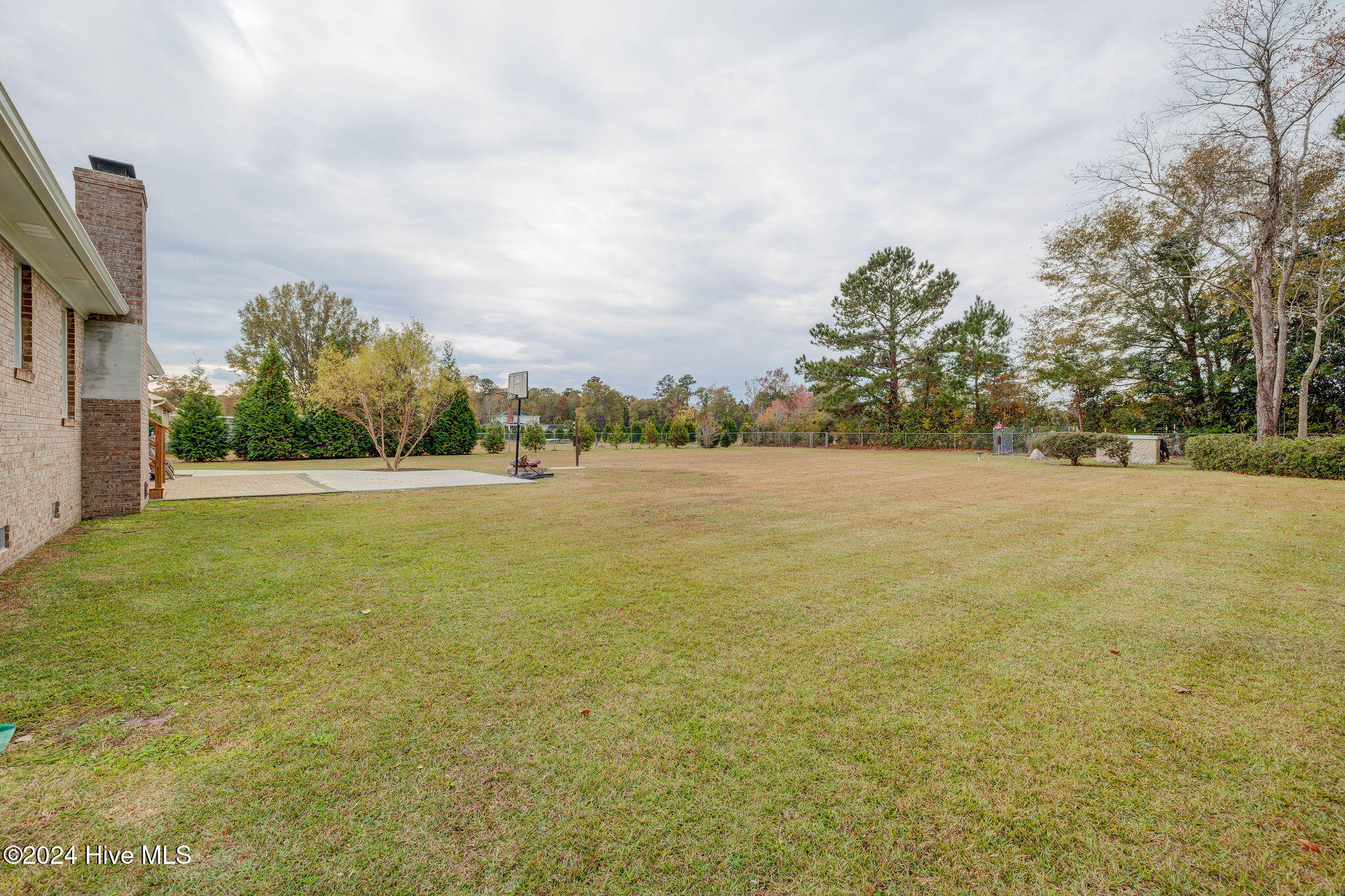 Spacious backyard with a lawn, paved patio, basketball hoop, and trees along a fence.