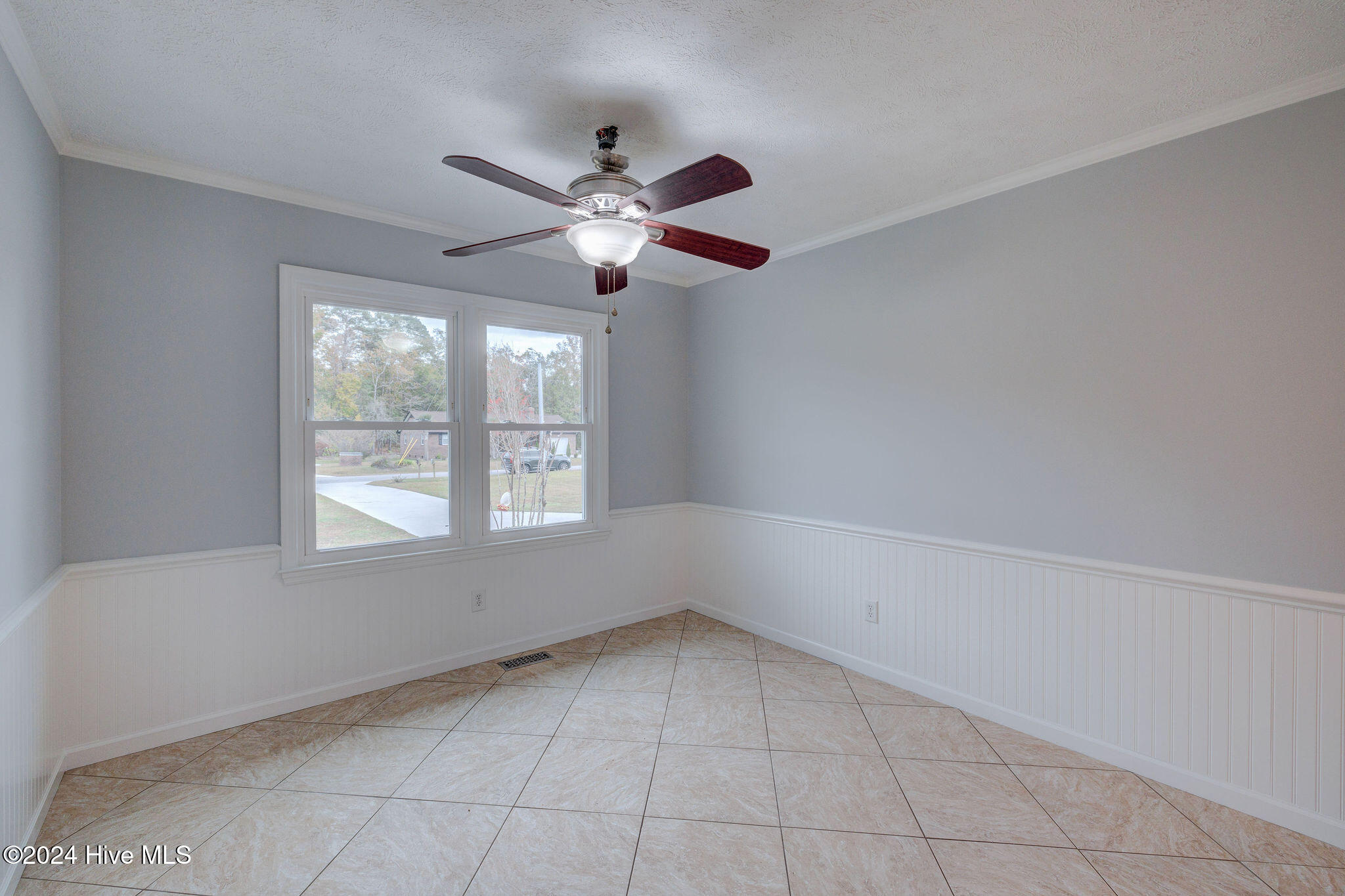 Empty room with beige tile floors, white wainscoting, two windows, and a dark wood ceiling fan.