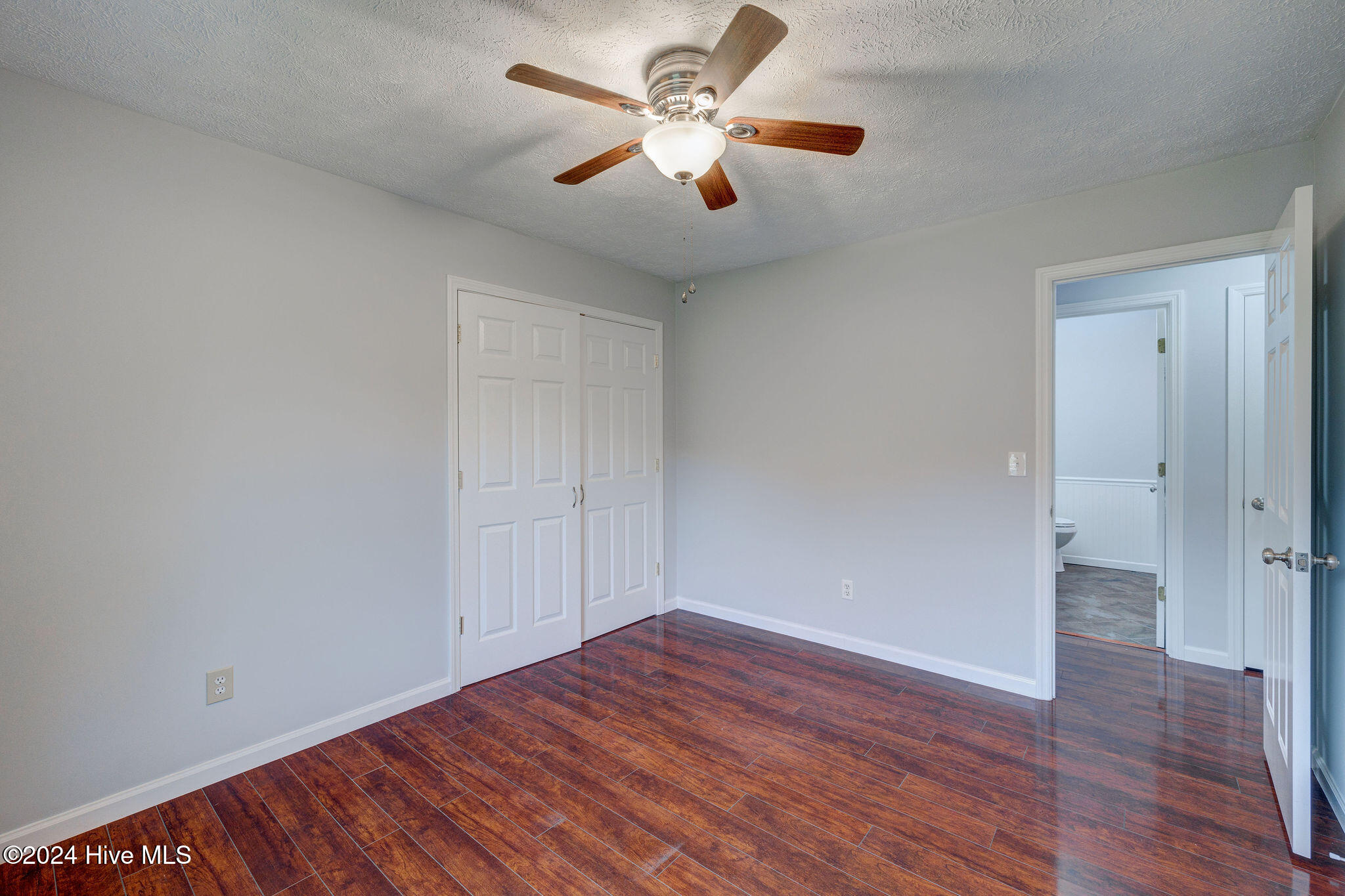 Empty bedroom with a wooden-blade ceiling fan, glossy plank flooring, and double closet doors.