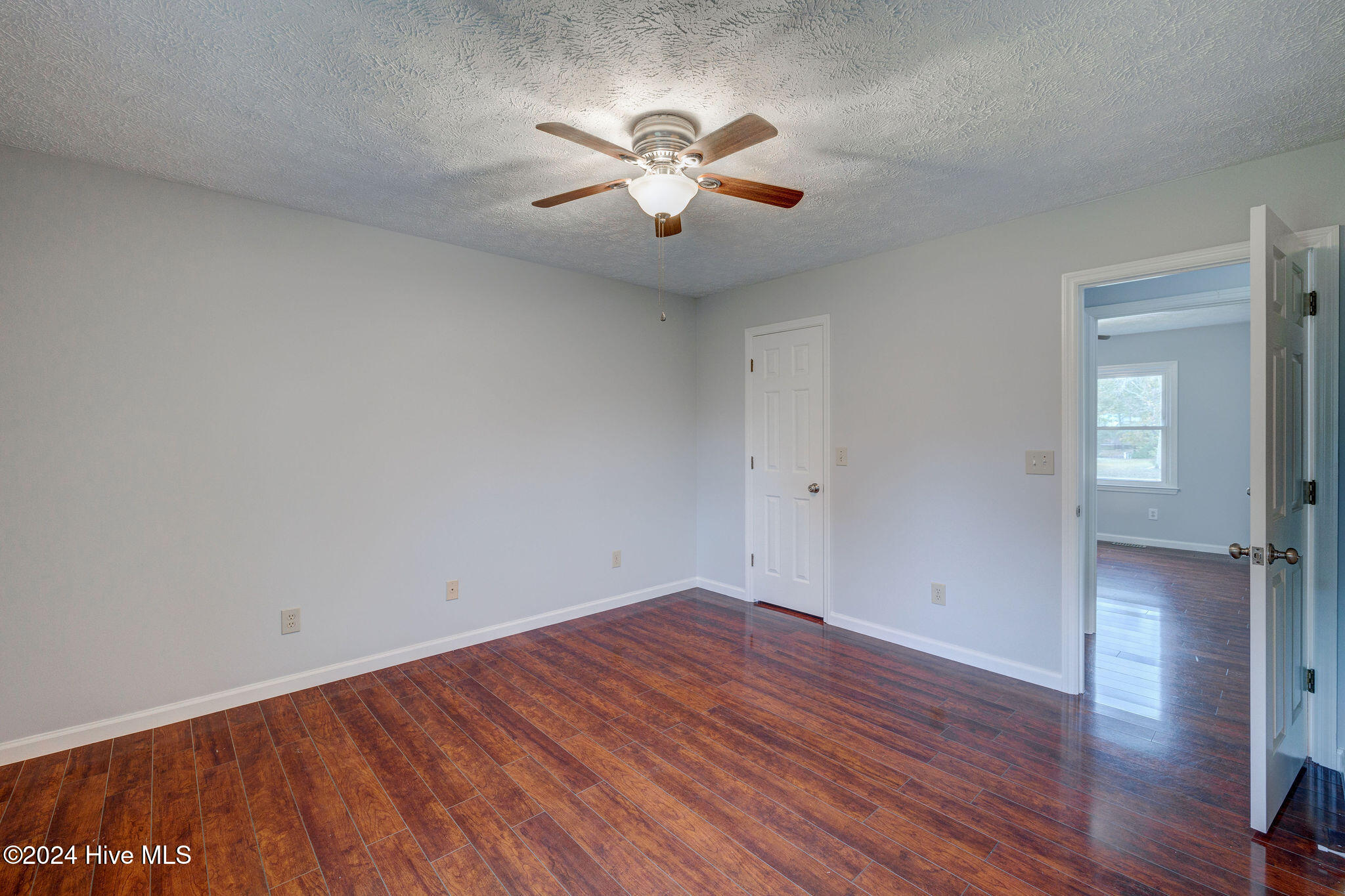 Vacant bedroom with reddish-brown flooring, a ceiling fan, and an open doorway to an adjoining room.