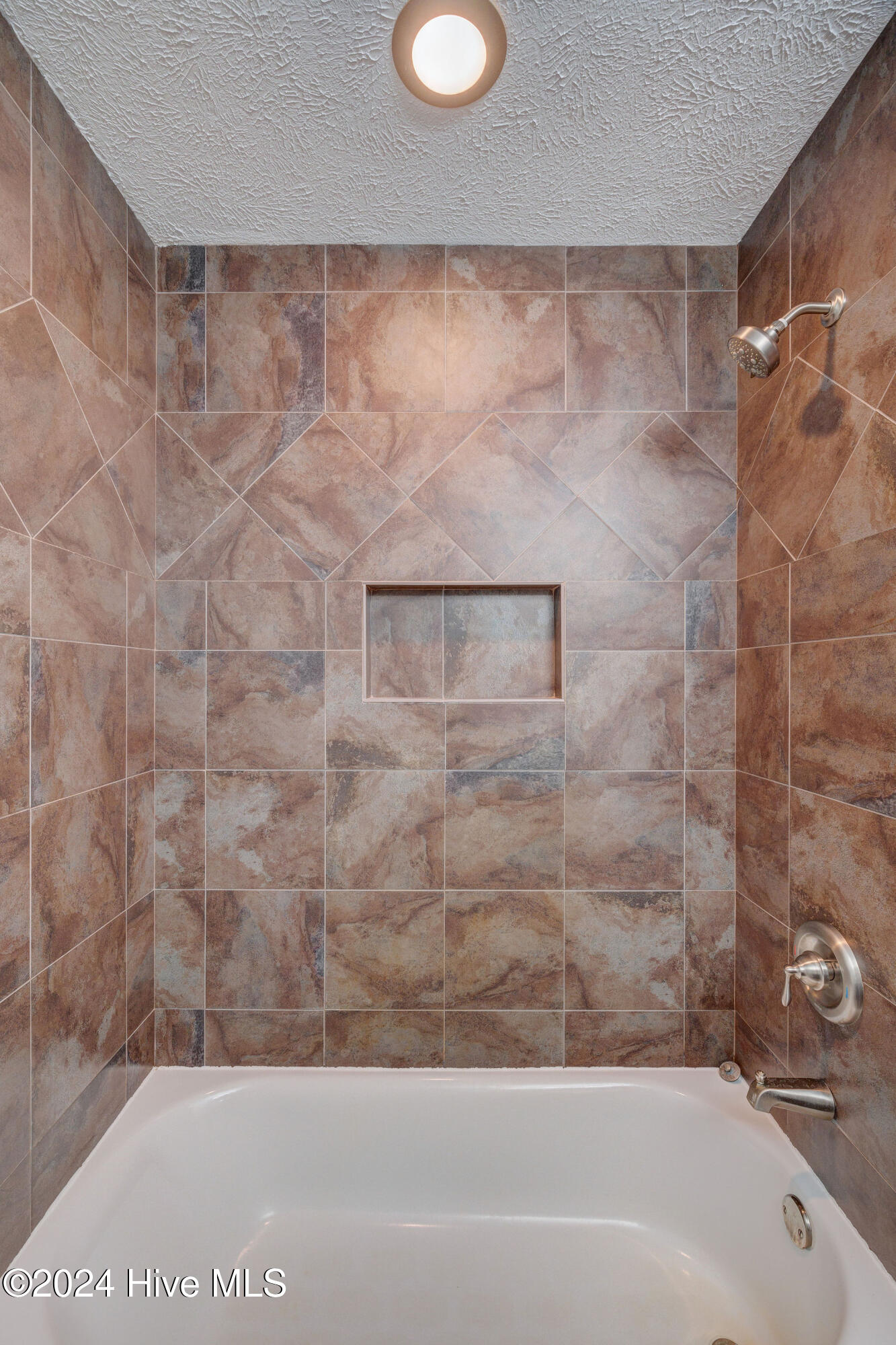 White bathtub with mottled brown tile shower walls, built-in niche, and brushed metal fixtures.