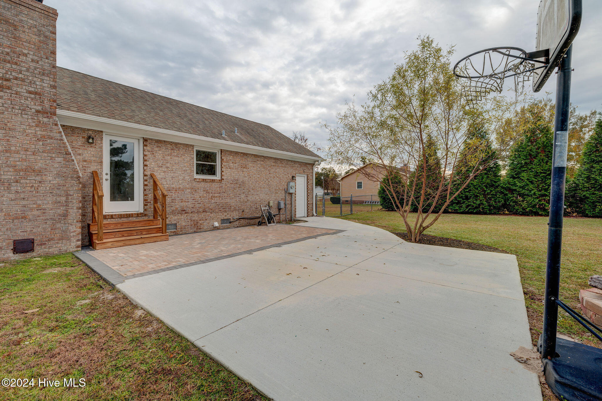 Brick home’s backyard with a paved patio, basketball hoop, fenced lawn, and leafy tree.