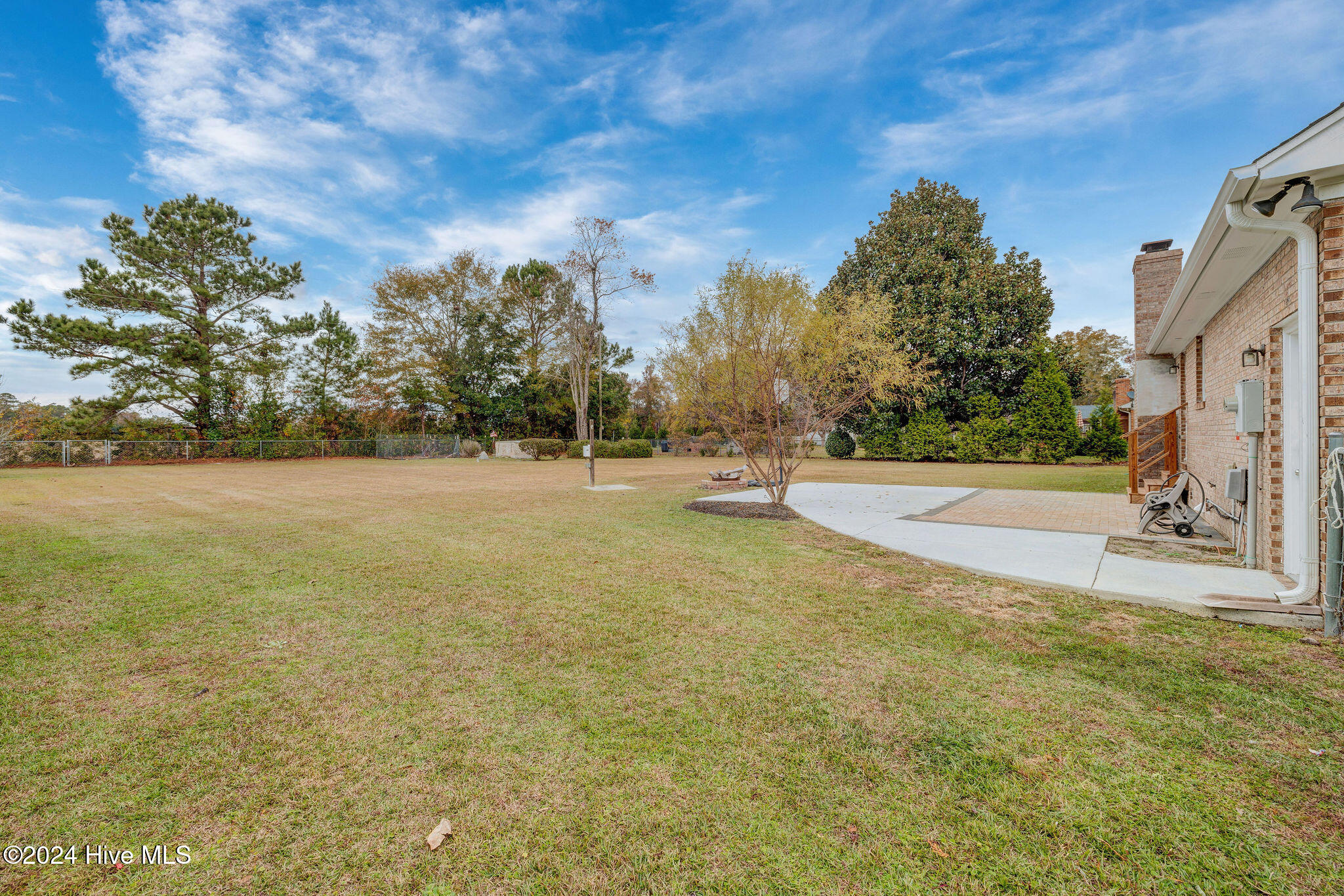 Grassy backyard with a concrete patio, brick-paved area, mature trees, and fenced lawn.