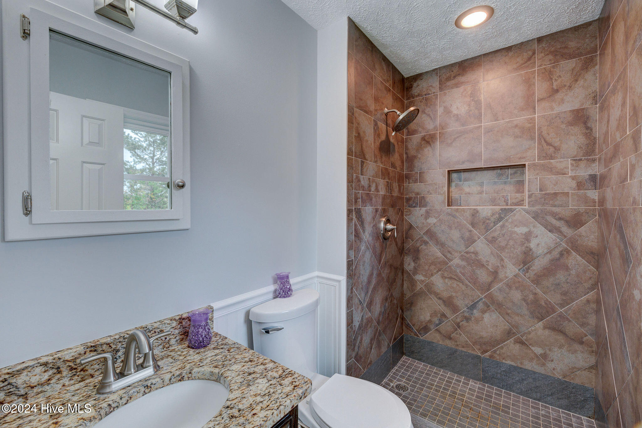 Bathroom with a brown-tiled walk-in shower, recessed shelf, granite vanity, and mirrored cabinet.