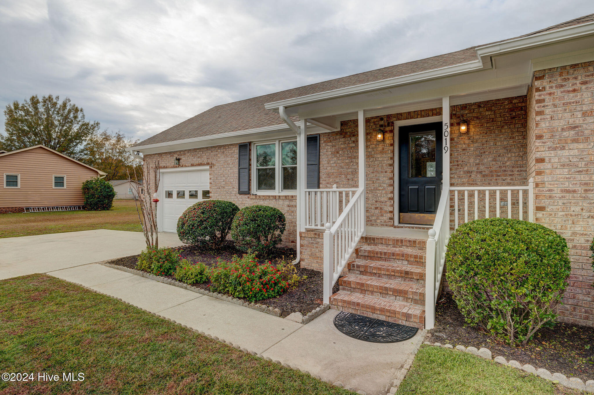 Brick home with a covered porch, brick steps, white garage door, and landscaped shrubs along the driveway.