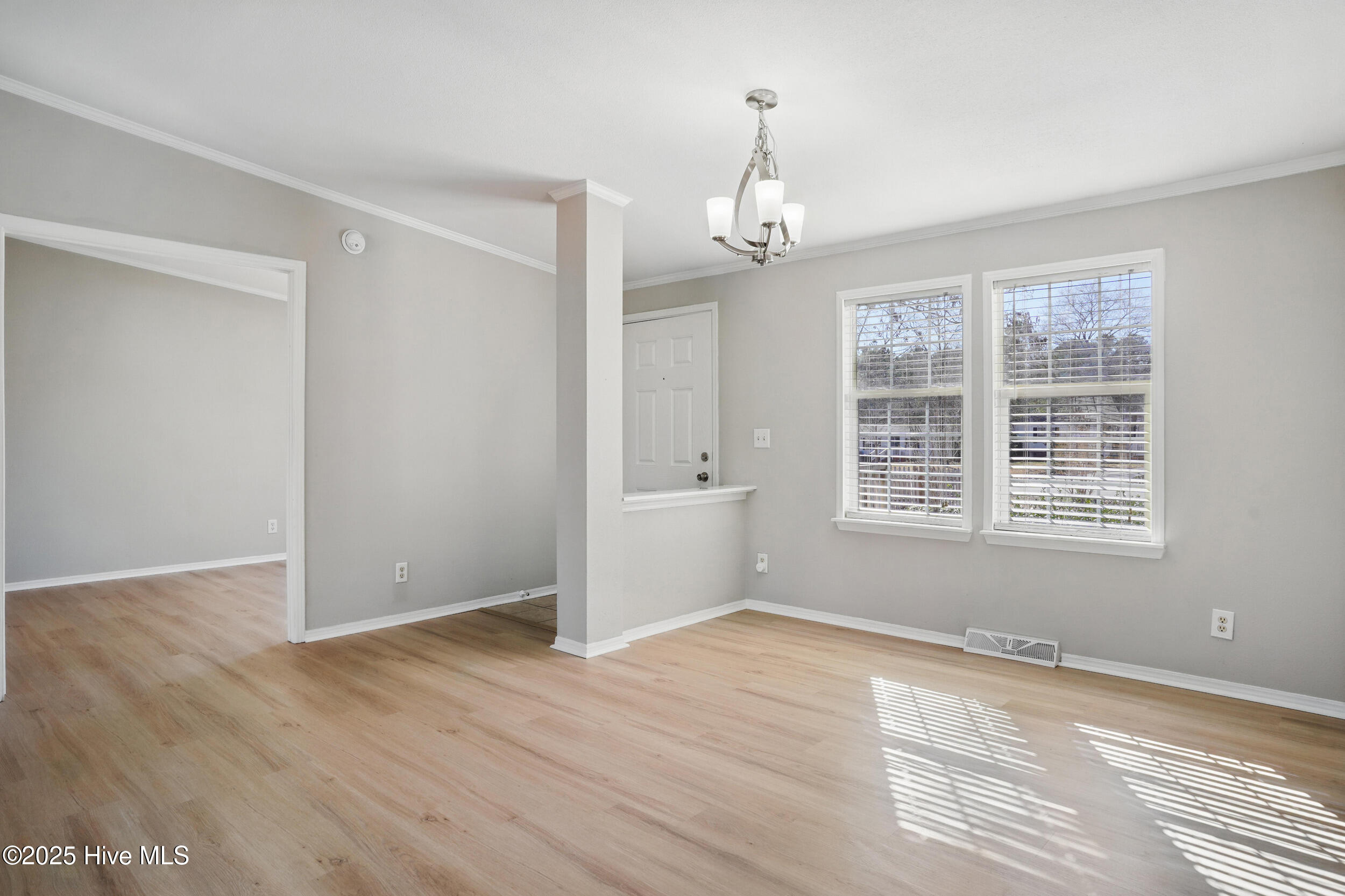 Empty dining area with pale wood floors, a chandelier, and sunlight through two blind-covered windows.