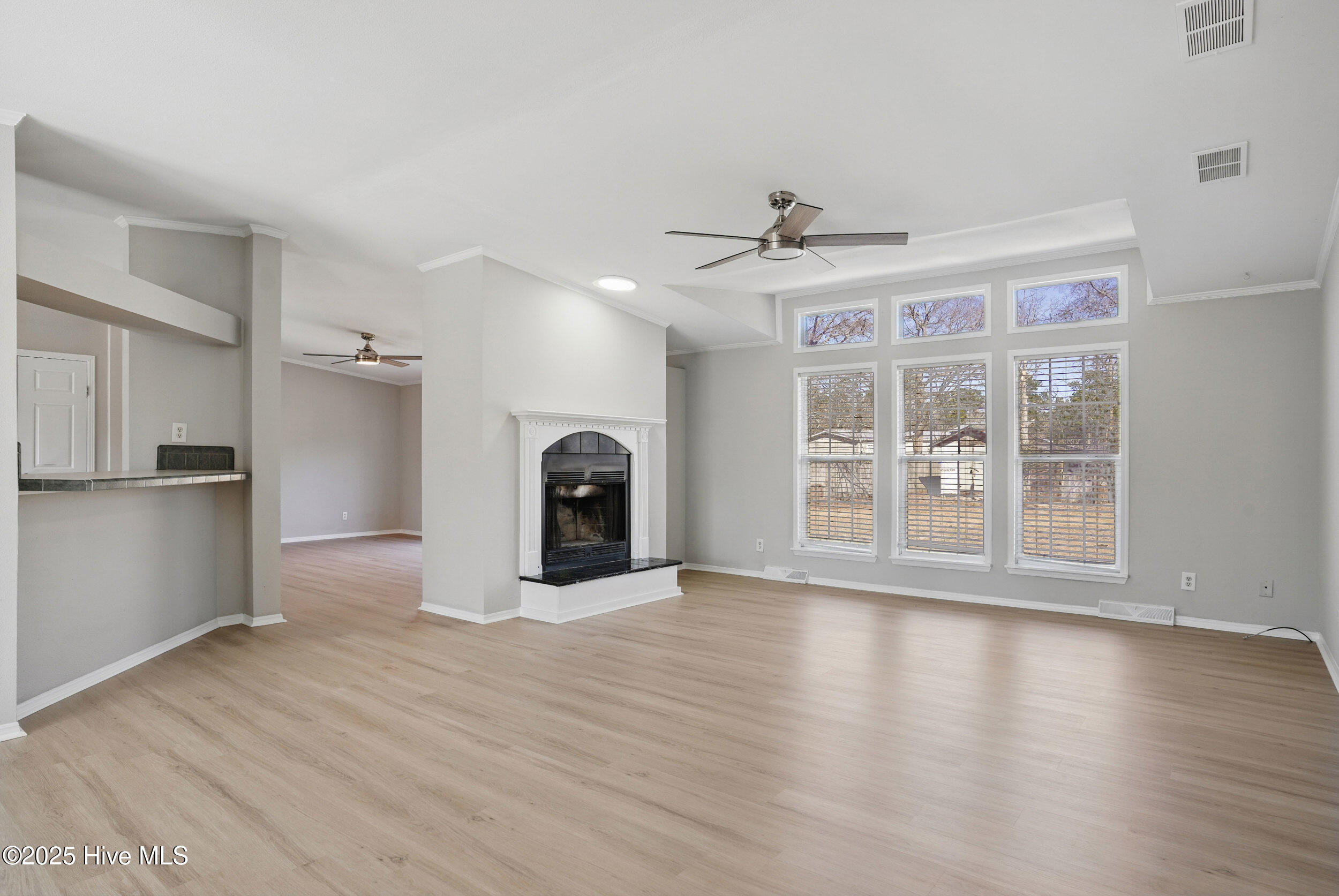 Vacant living room with a fireplace, light wood-look floors, and tall windows with blinds.