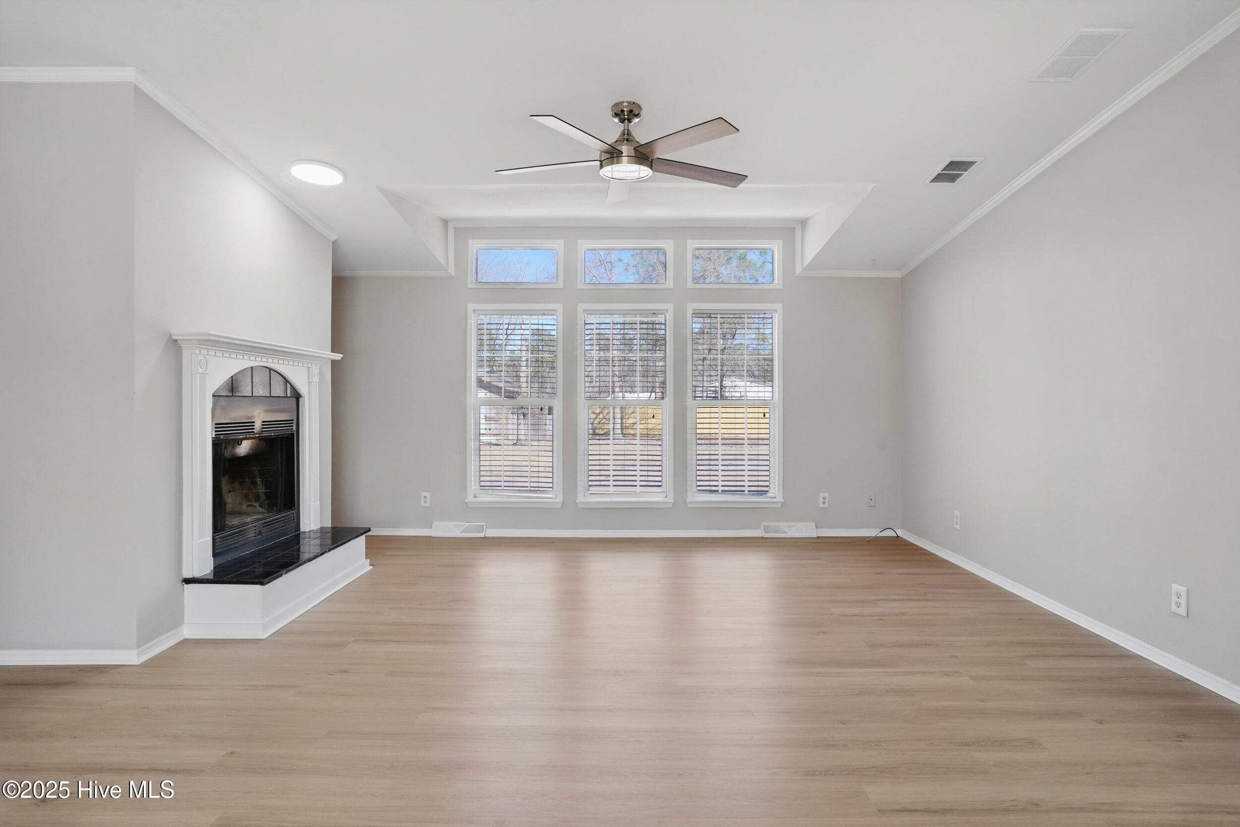 Vacant living room with a fireplace, tall windows, light wood floors, and ceiling fan.
