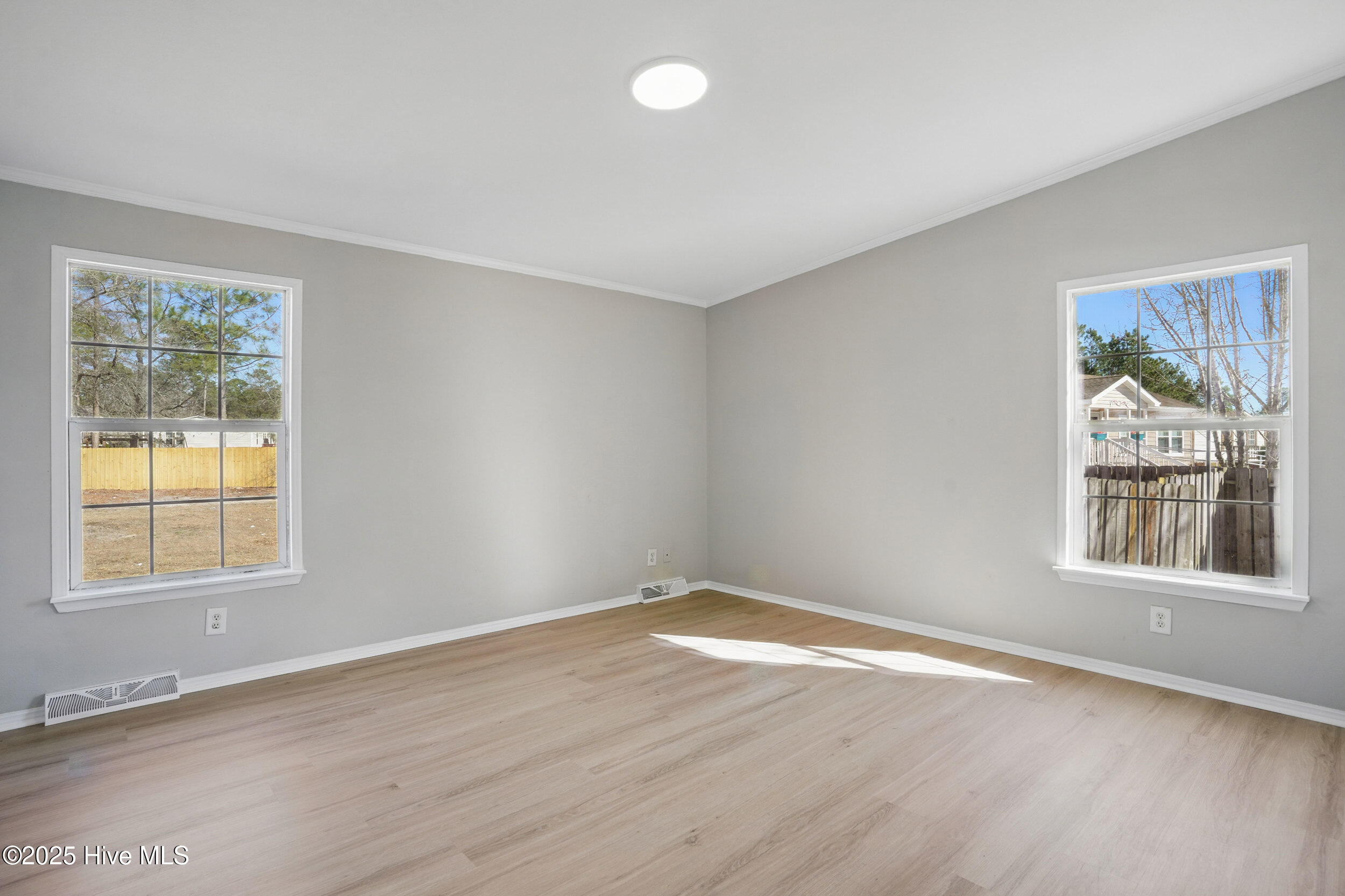 Empty room with light wood floors, gray walls, and two windows overlooking a fenced yard.