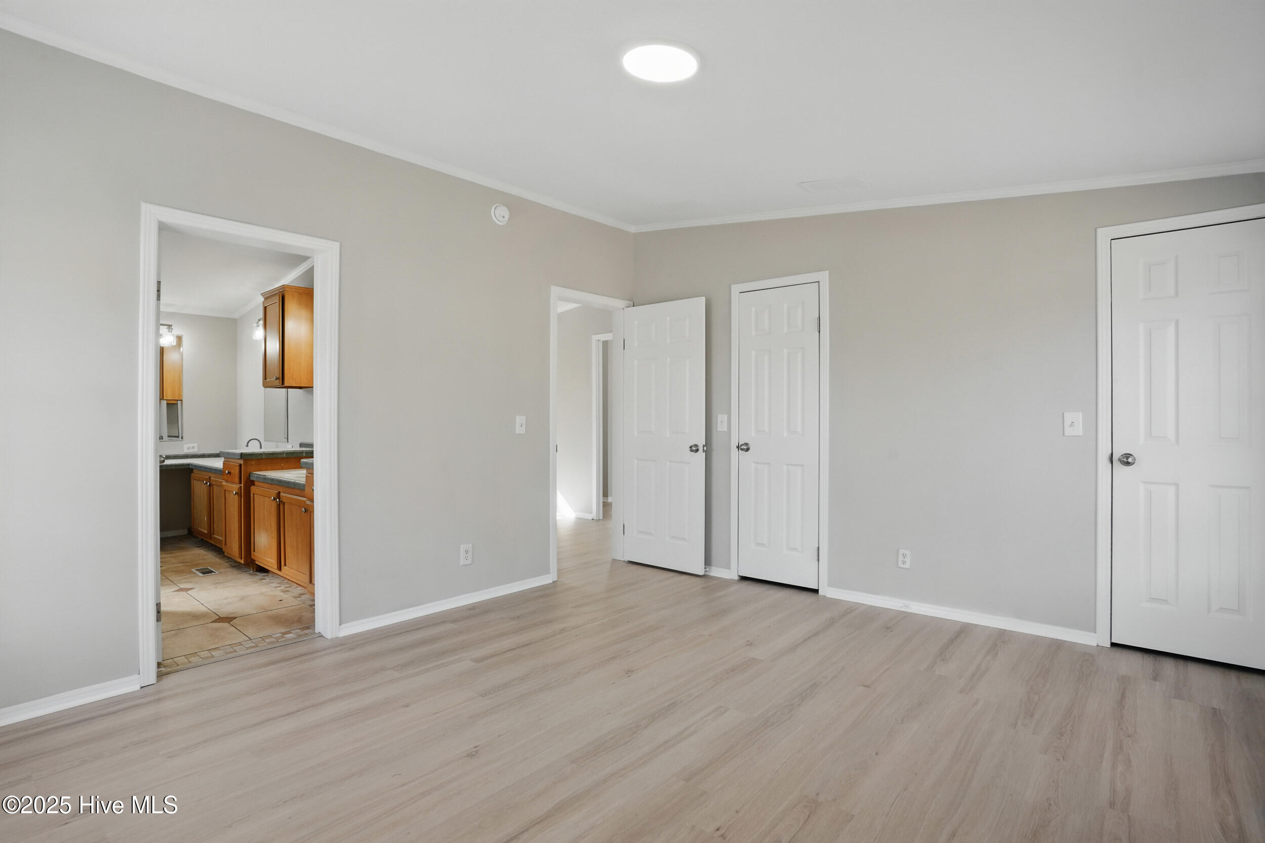 Empty room with light wood-look floors and gray walls, opening to a kitchen with wood cabinets.