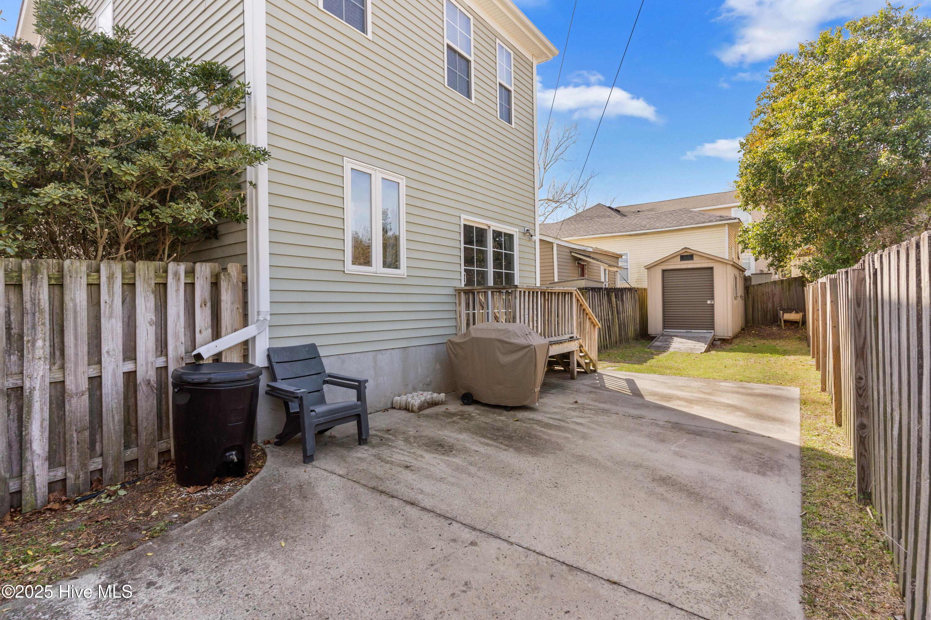 Fenced backyard with a concrete patio, small deck, covered grill, and storage shed beside a two-story house.