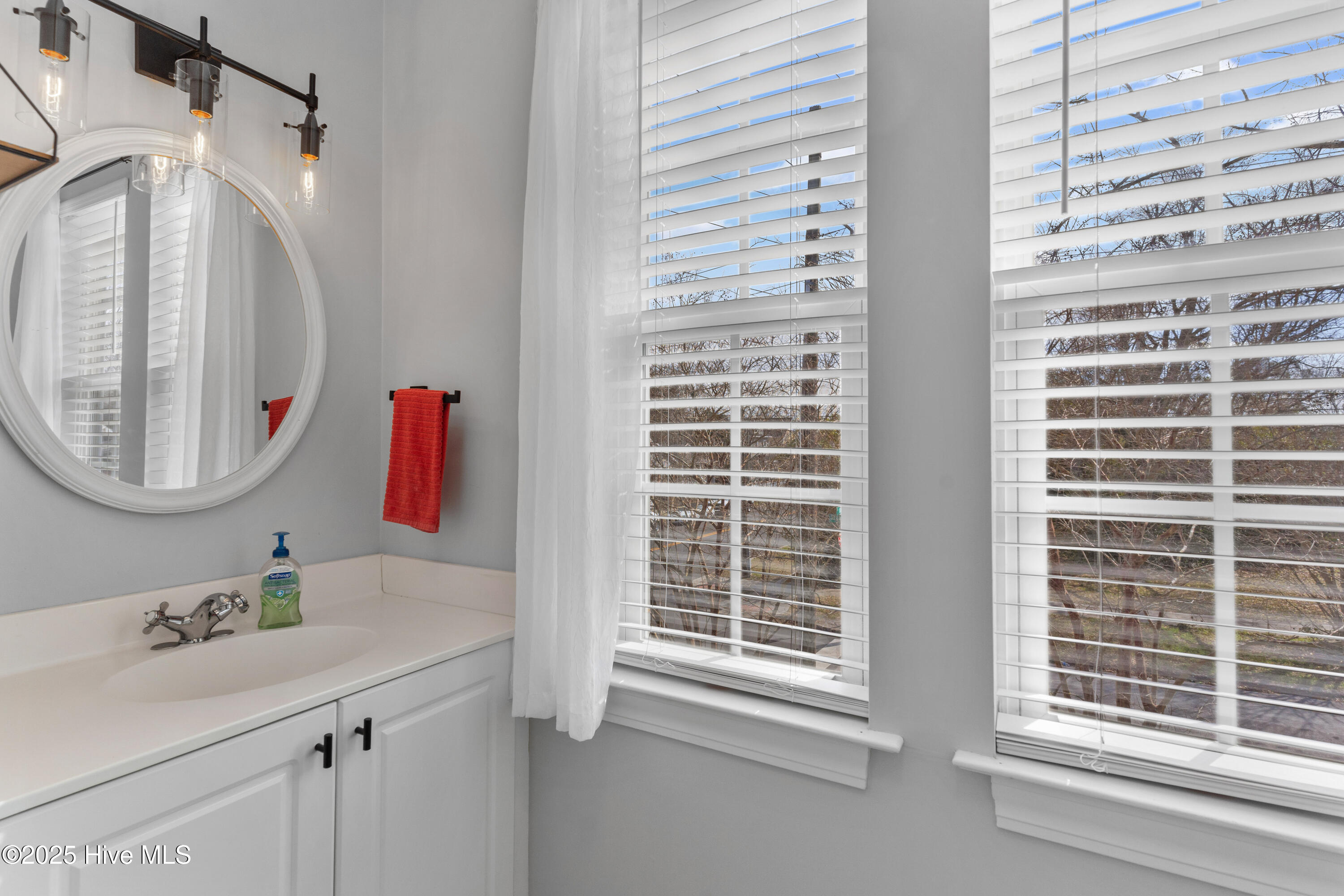 Bathroom vanity with round mirror and red hand towel beside two large windows with white blinds.