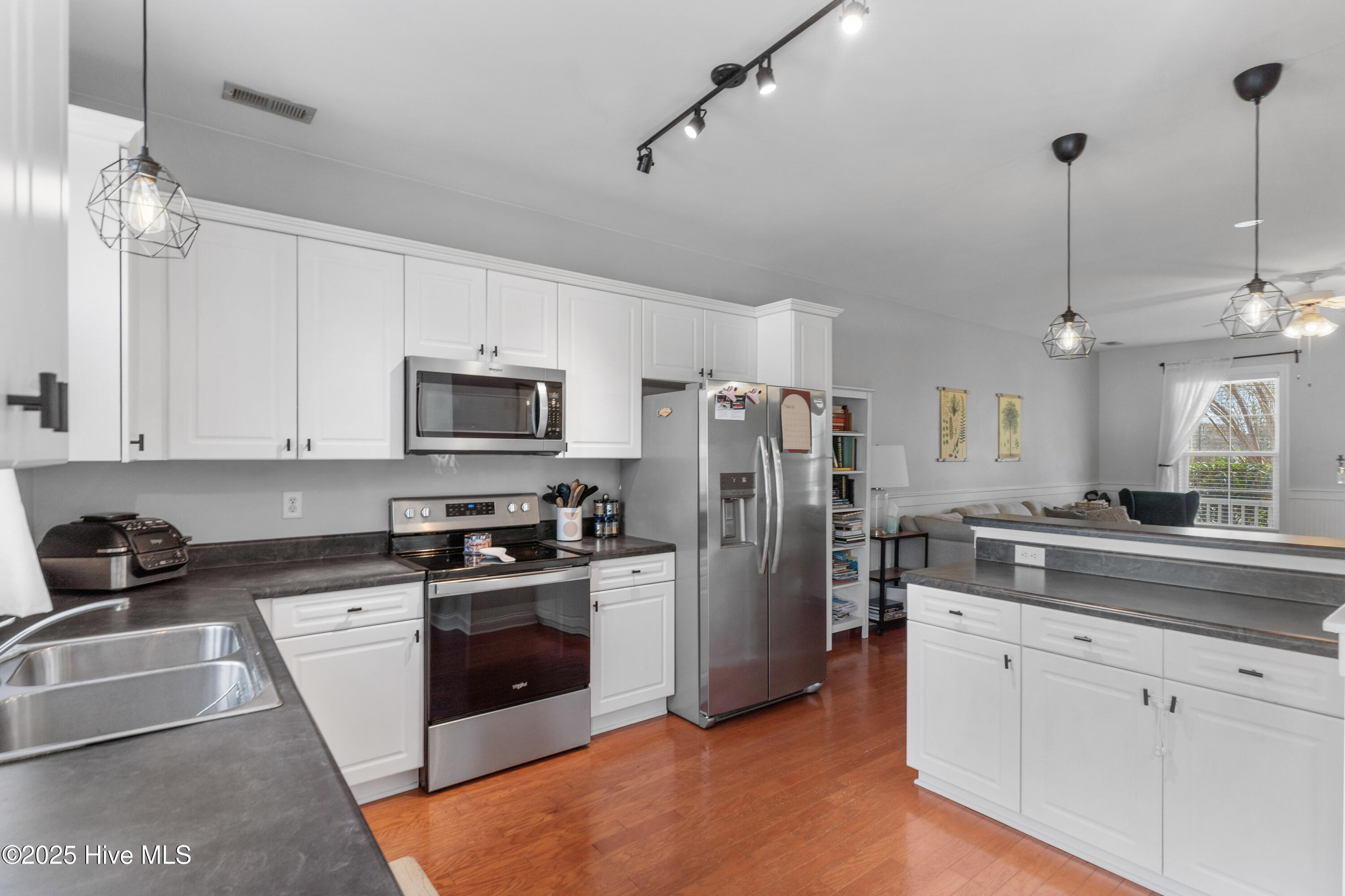 Kitchen with white cabinets, stainless steel appliances, dark countertops, and hardwood floors opening to a living area.
