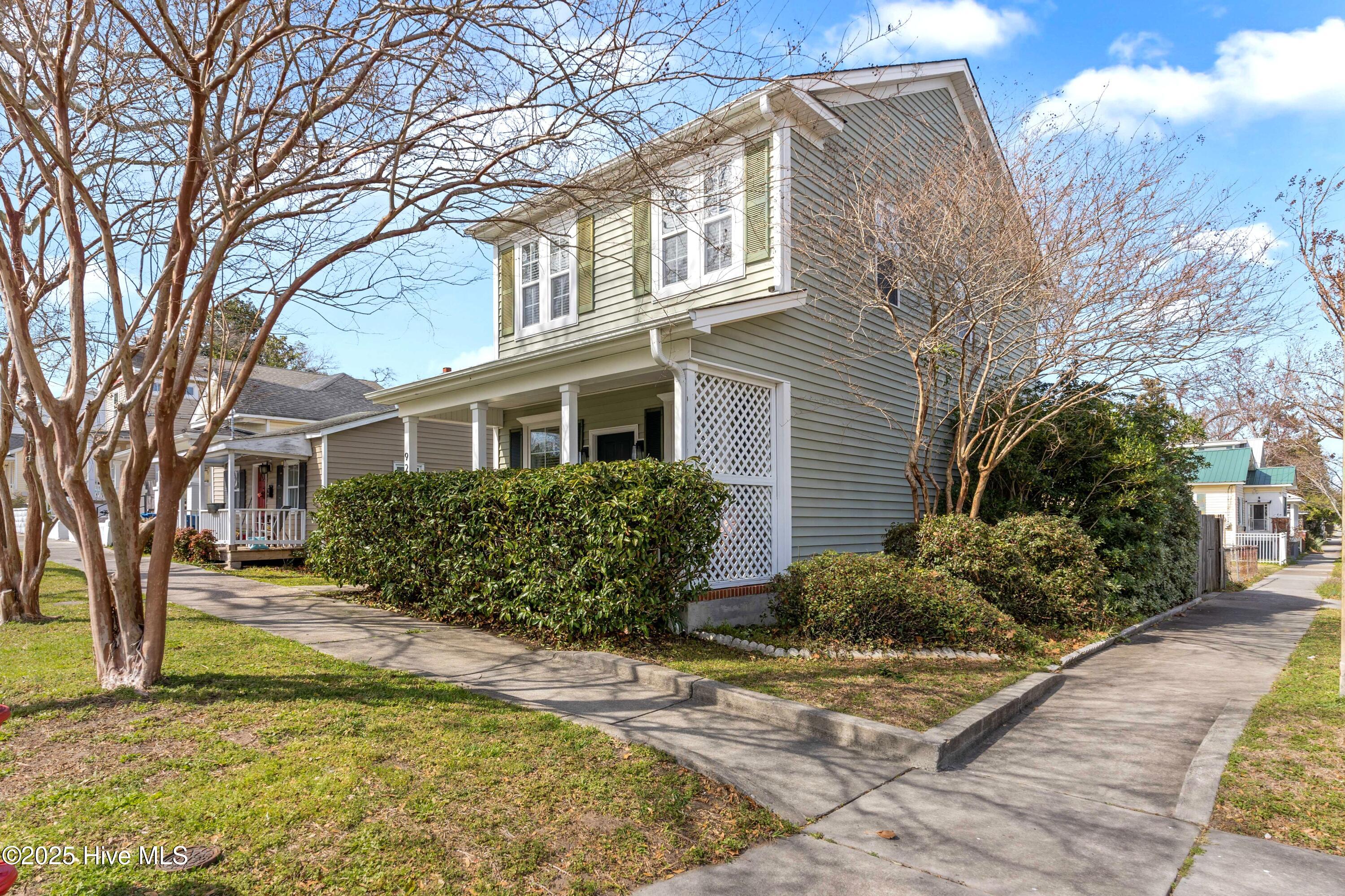 Two-story gray-green house with green shutters, a covered porch, and hedges along the sidewalk.
