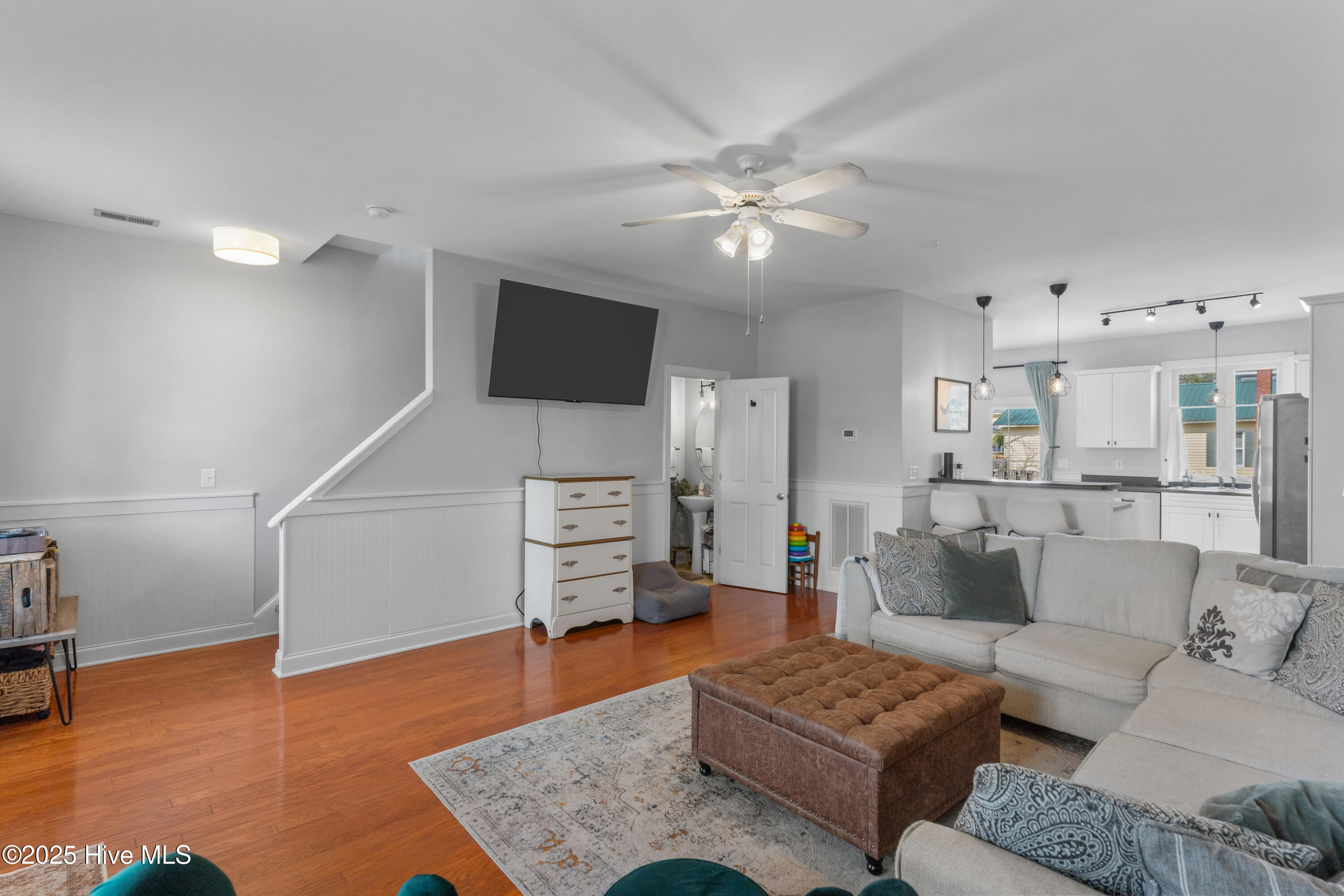 Living room with a gray sectional, tufted ottoman, hardwood floors, and an open kitchen.