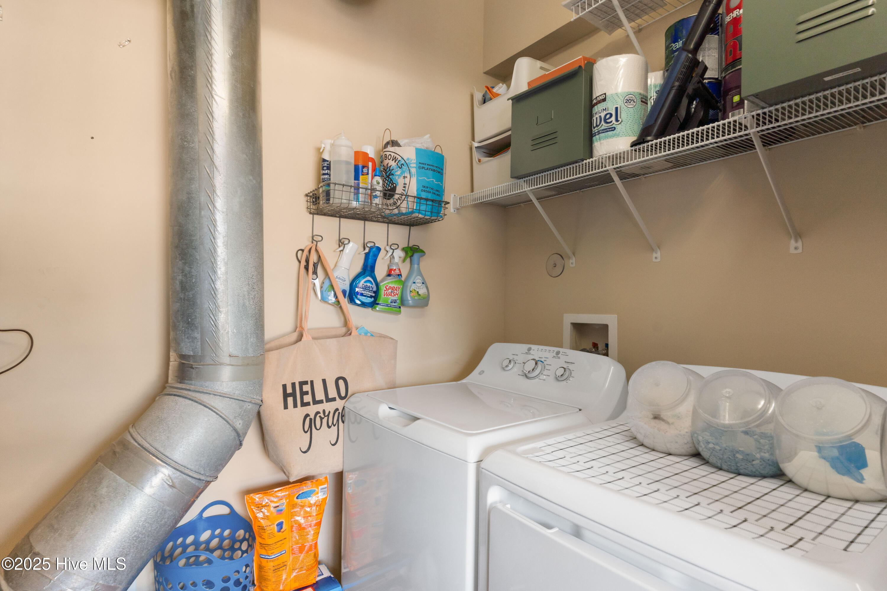 Laundry room with washer and dryer, wire shelves, cleaning supplies, and a prominent metal vent duct.