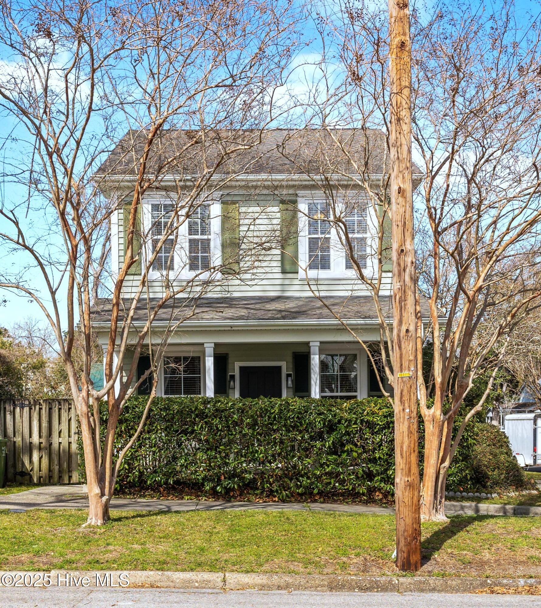Two-story white house with green shutters, partly screened by leafless trees and a tall utility pole.