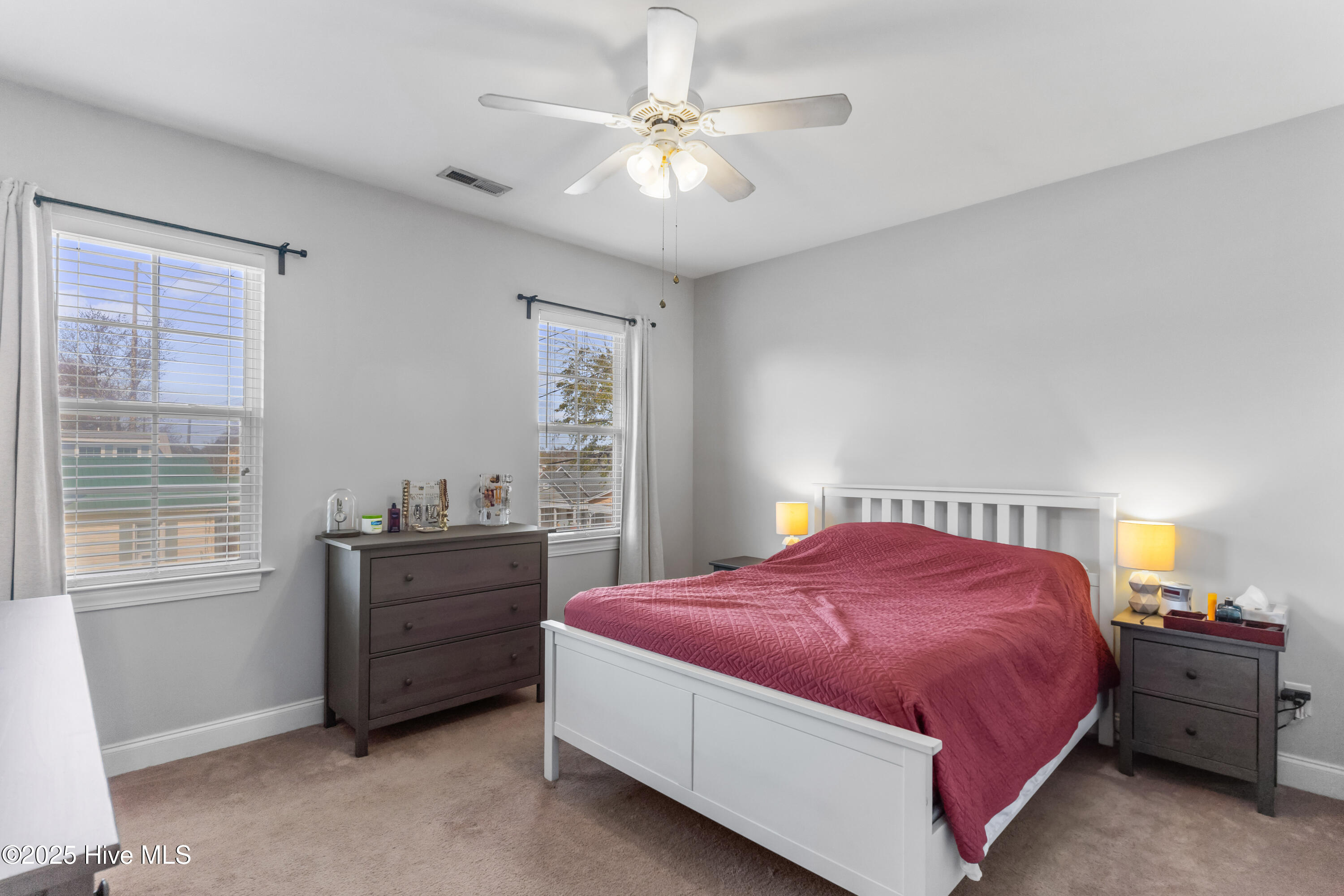 Bedroom with a white bed and rose-colored quilt, dark dressers, and two curtained windows.