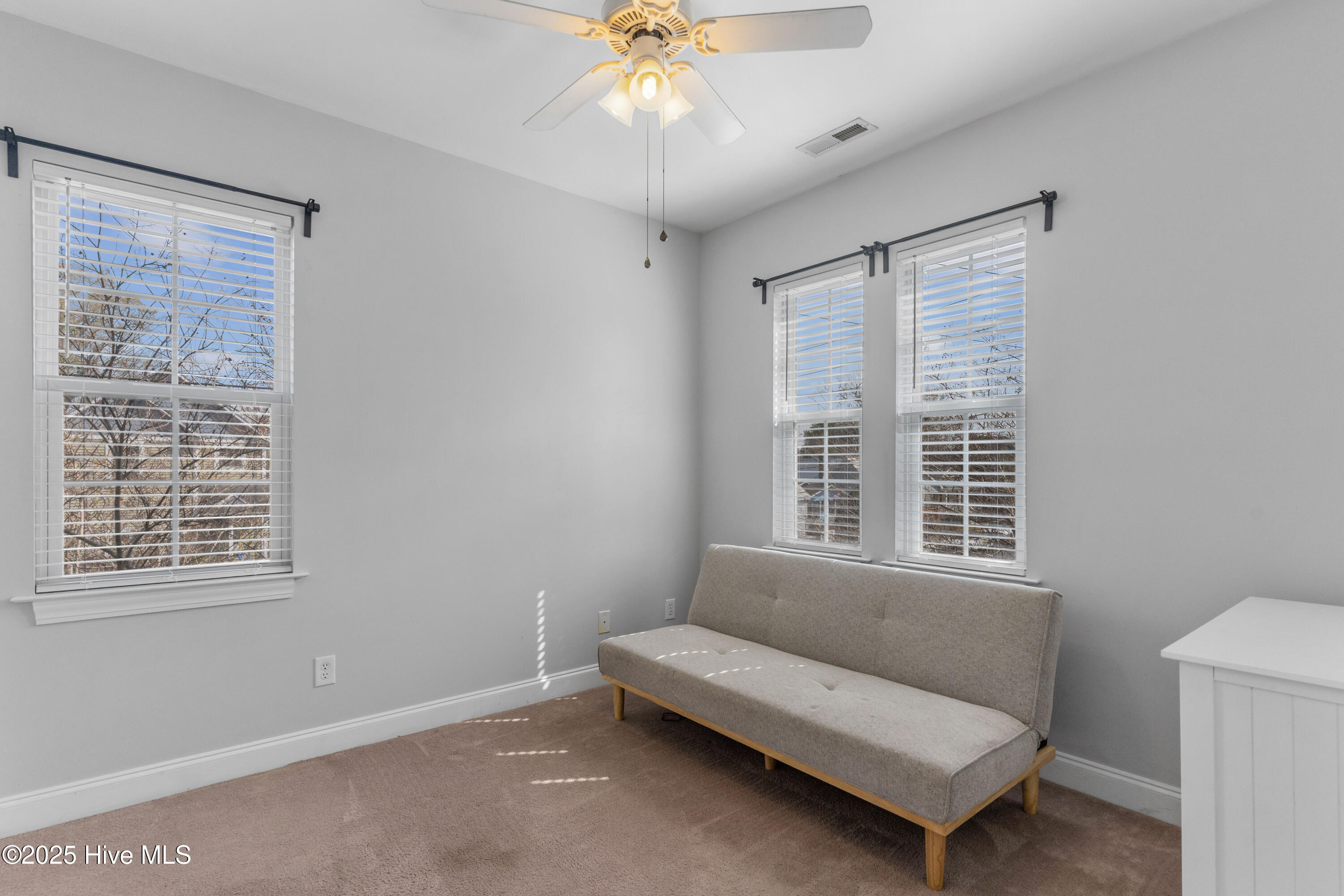 Bright room with a gray sofa, three blind-covered windows, beige carpet, and a ceiling fan.