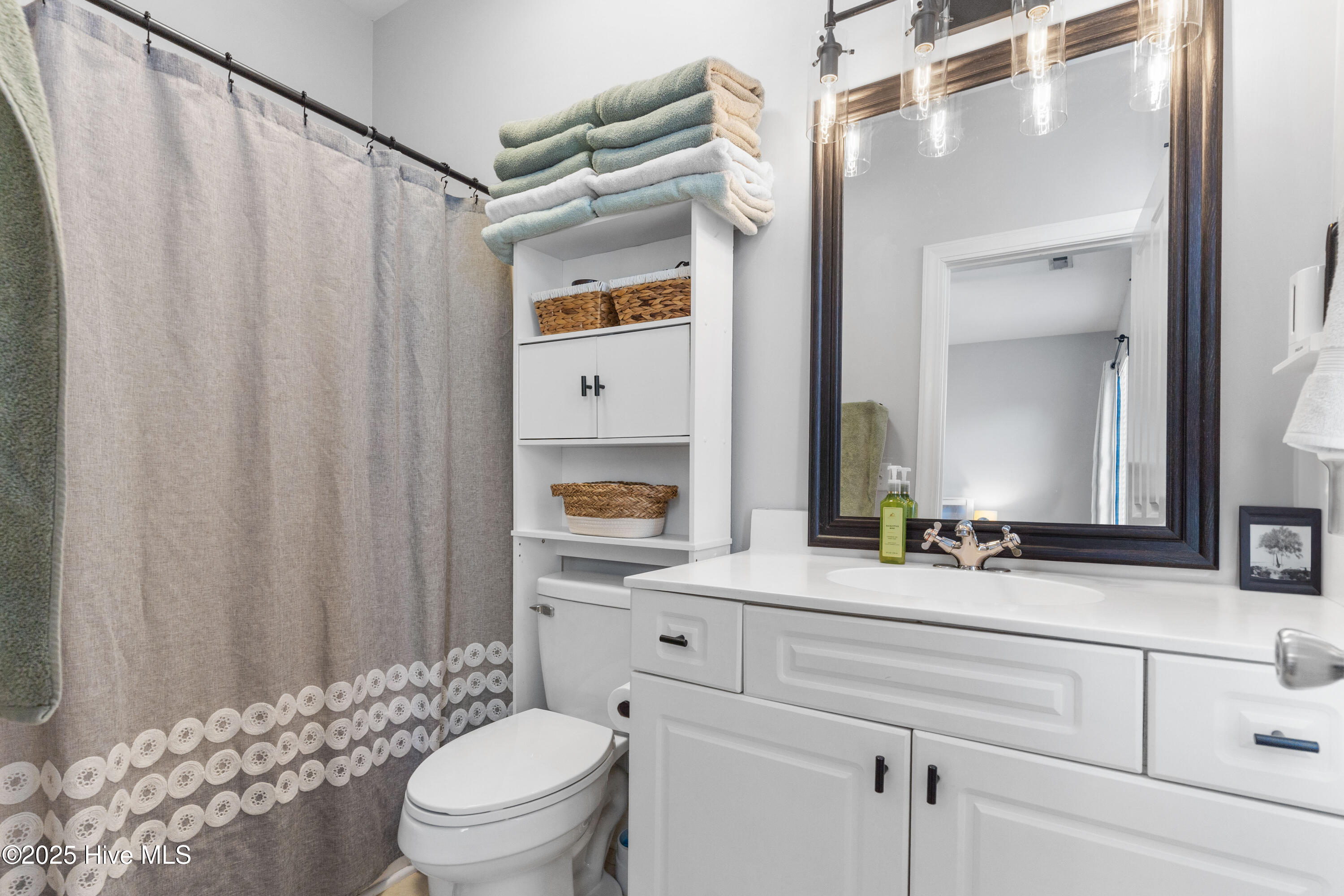 Bathroom with a white vanity, dark-framed mirror, lace-trimmed shower curtain, and shelves of folded towels.