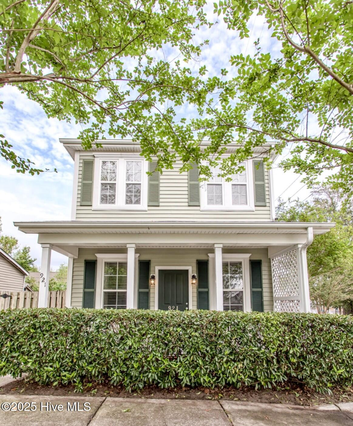 Two-story gray house with a white porch, dark green shutters, and leafy branches overhead.