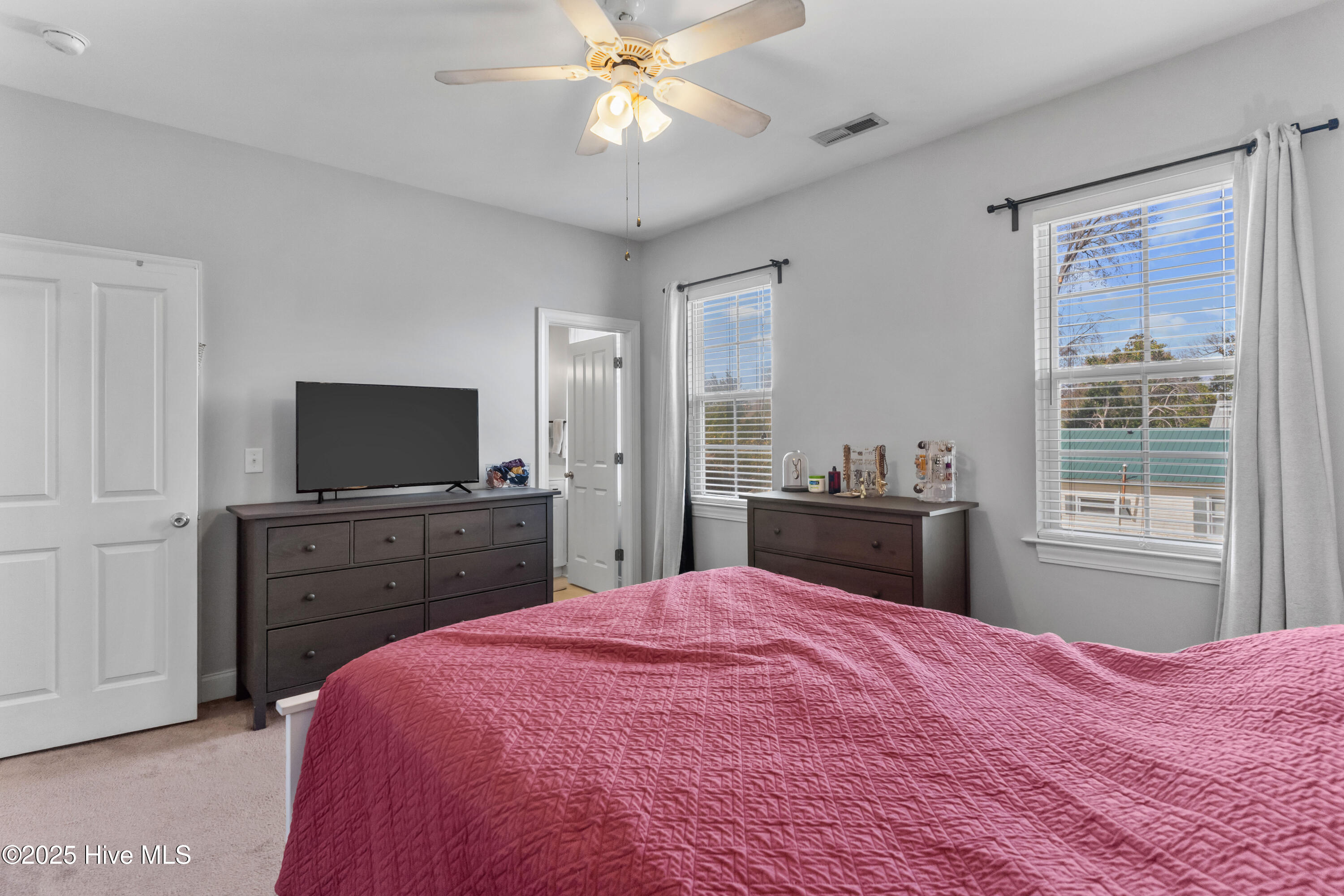 Bedroom with a pink quilted bed, dark dressers, and two curtained windows.