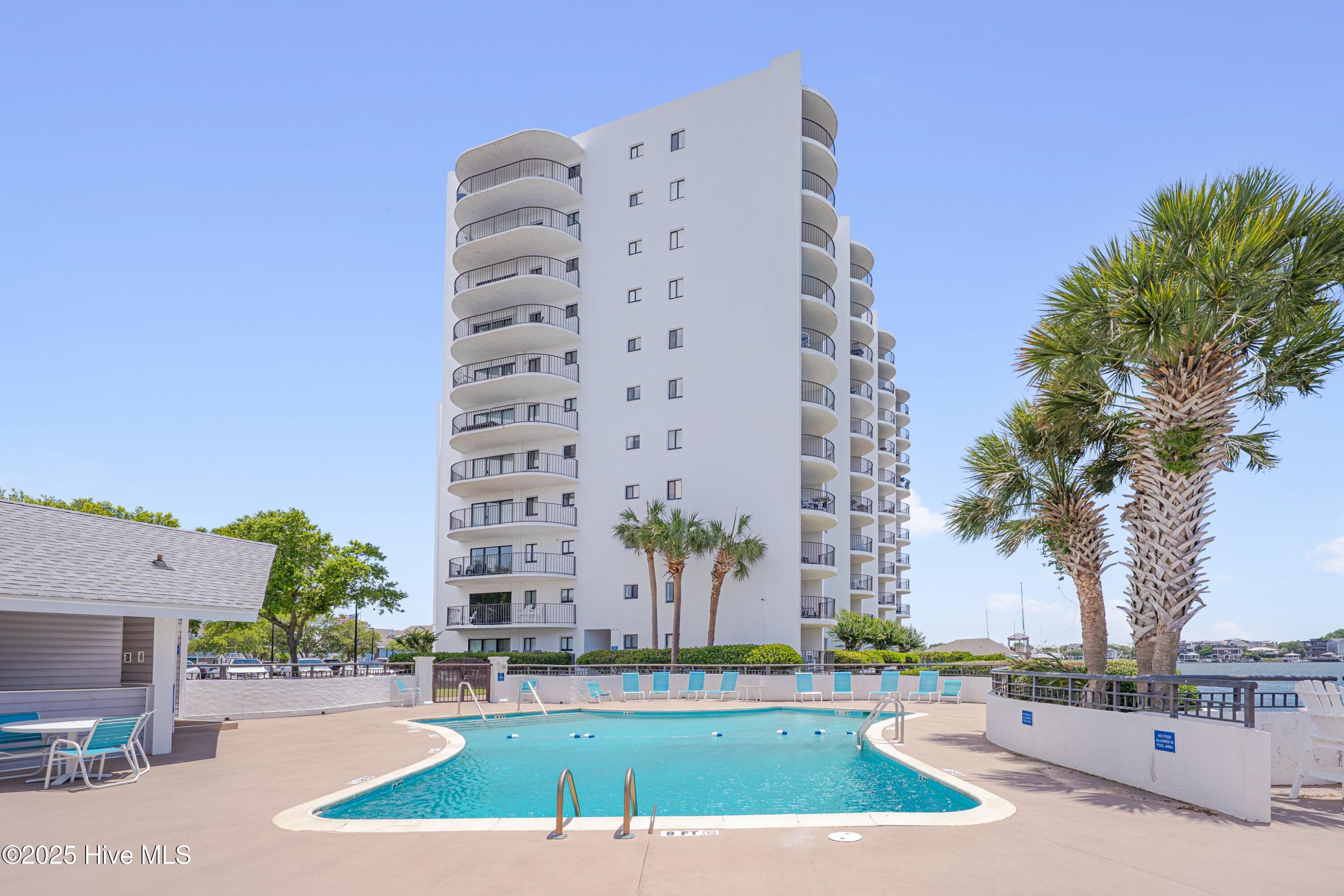 Condominium pool and deck with curved balconies, palm trees, and waterfront beyond.