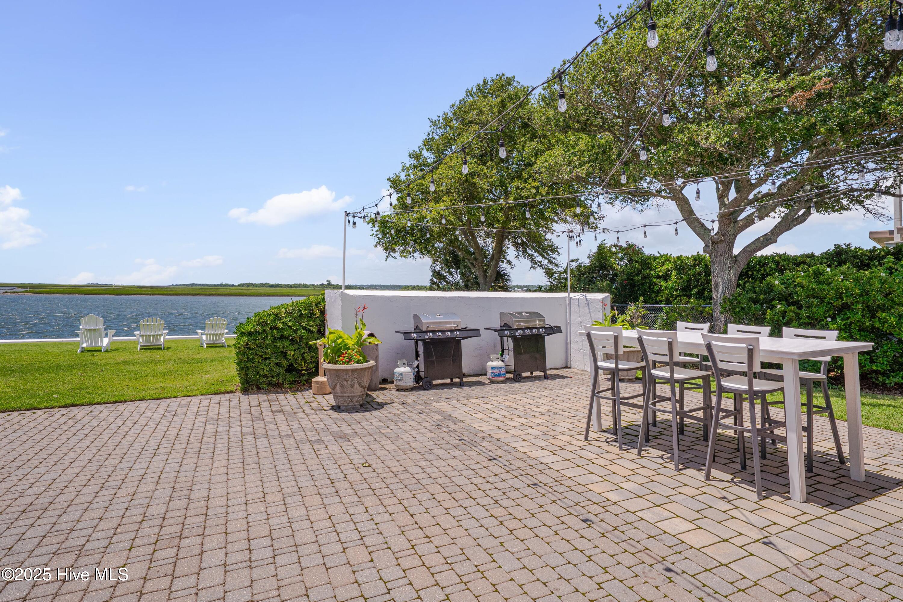 Waterfront patio with a paved dining area, string lights, two grills, and lawn chairs beside the water.