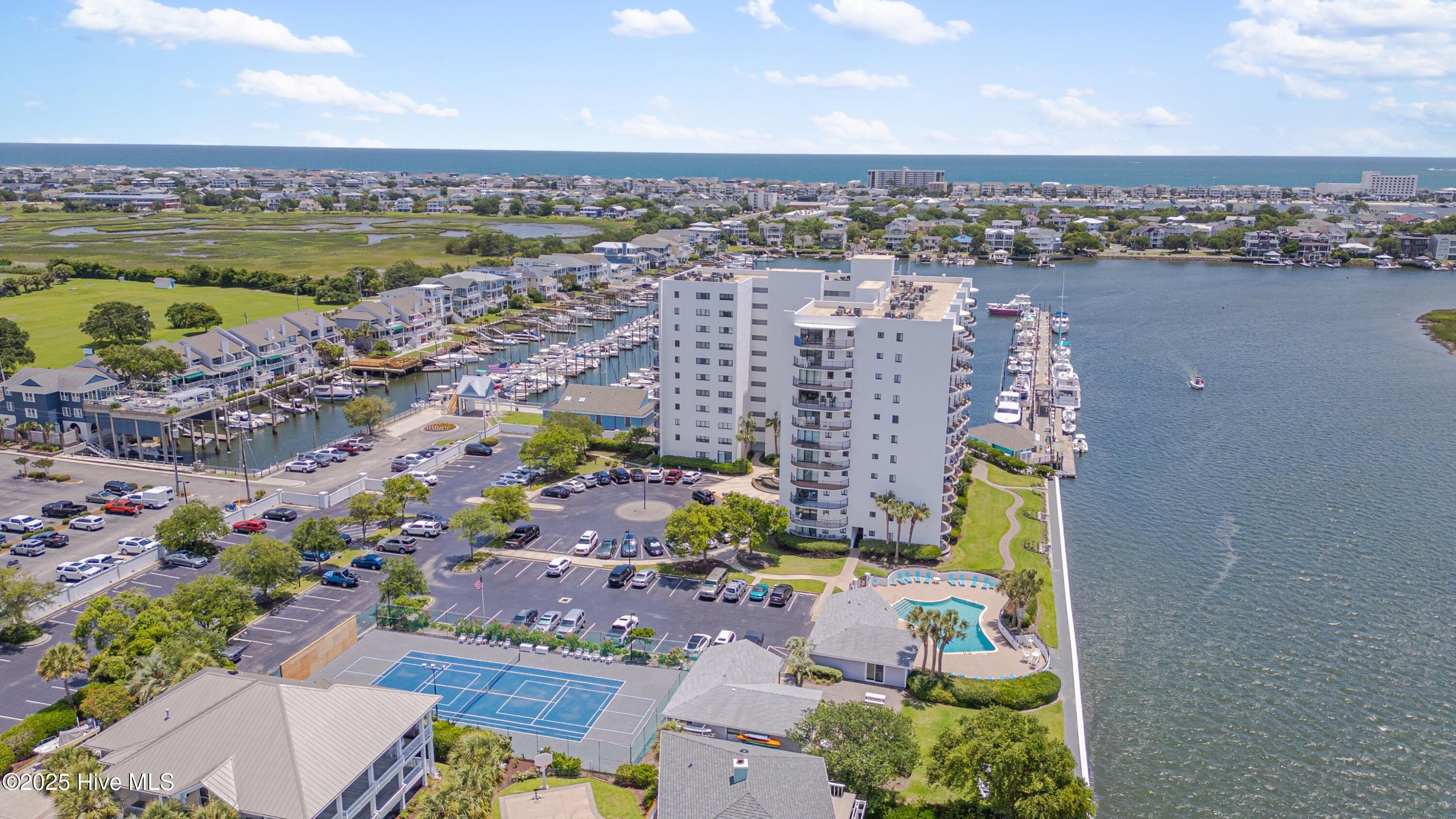 Waterfront condominium tower beside a marina, pool, and tennis courts, with the ocean beyond.