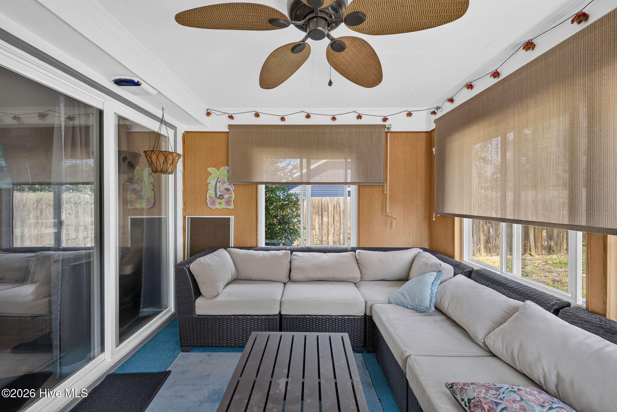 Enclosed sunroom with a gray sectional, blue flooring, wide windows, and a wicker-blade ceiling fan.