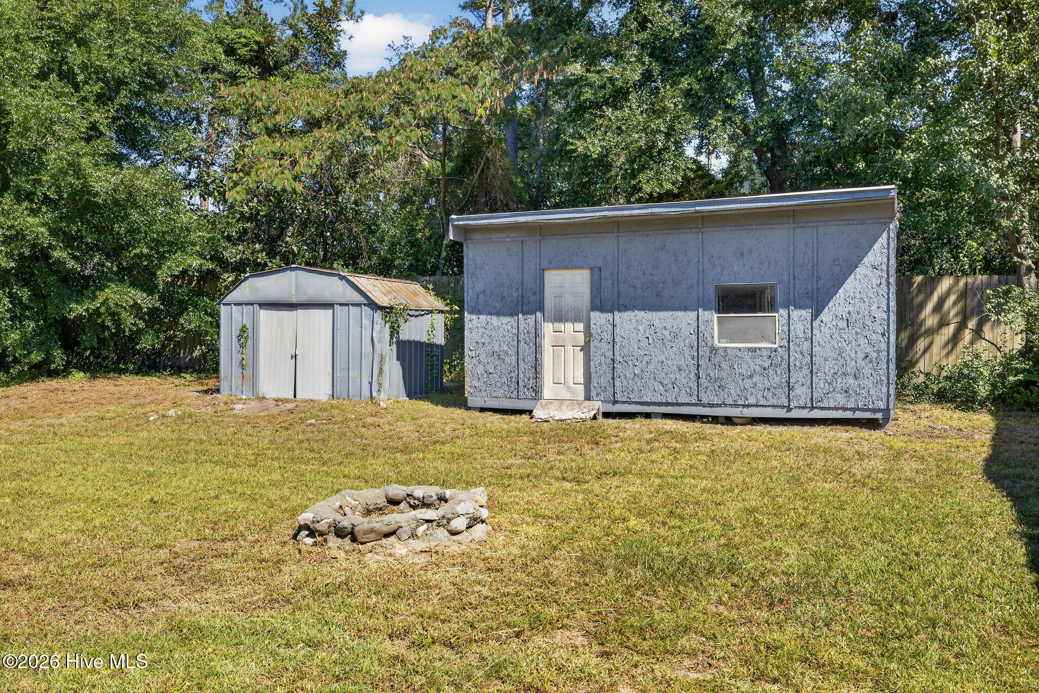 Gray detached outbuilding and metal shed beside a stone fire pit in a fenced yard.