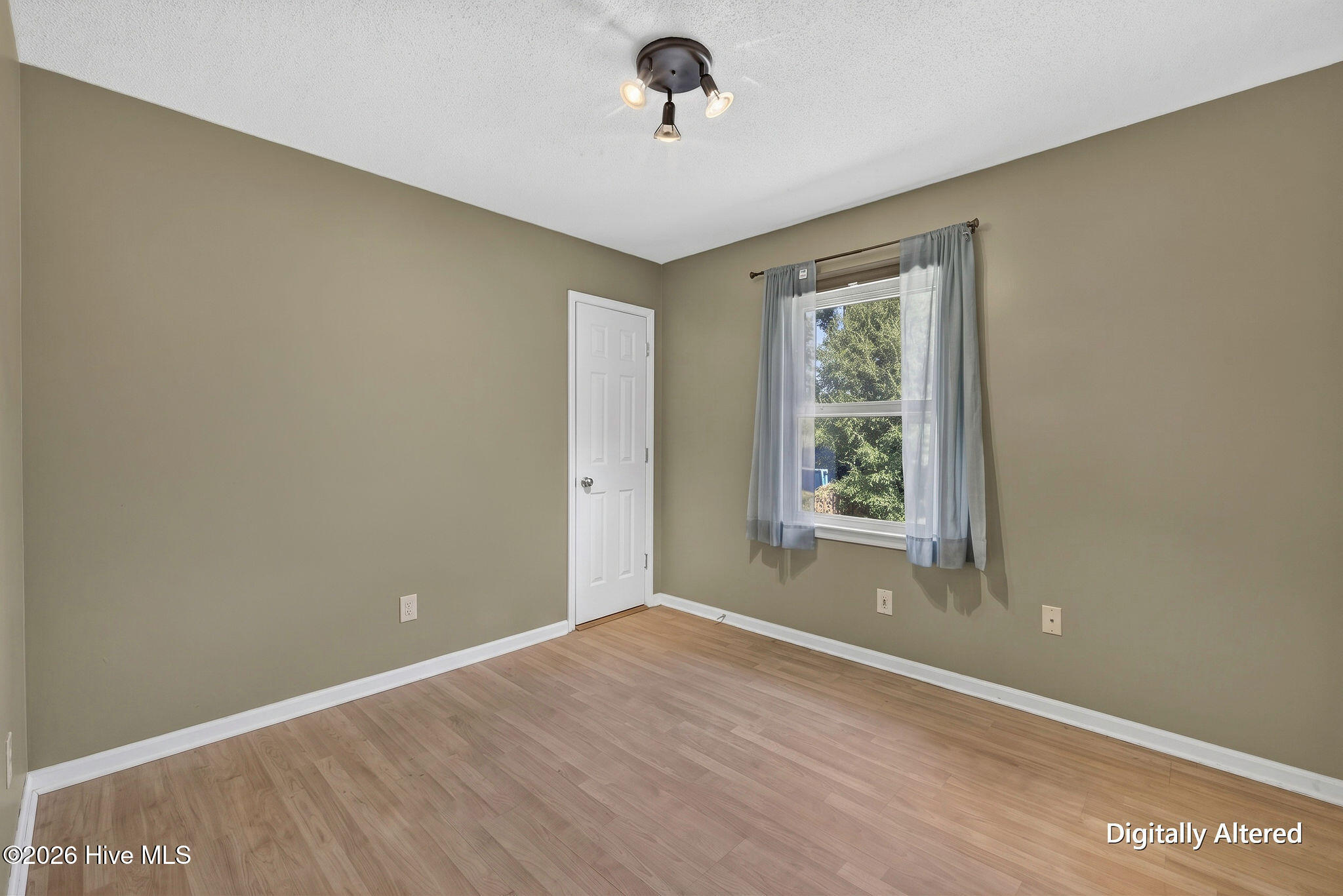 Empty bedroom with light wood flooring, taupe walls, and a curtained window.